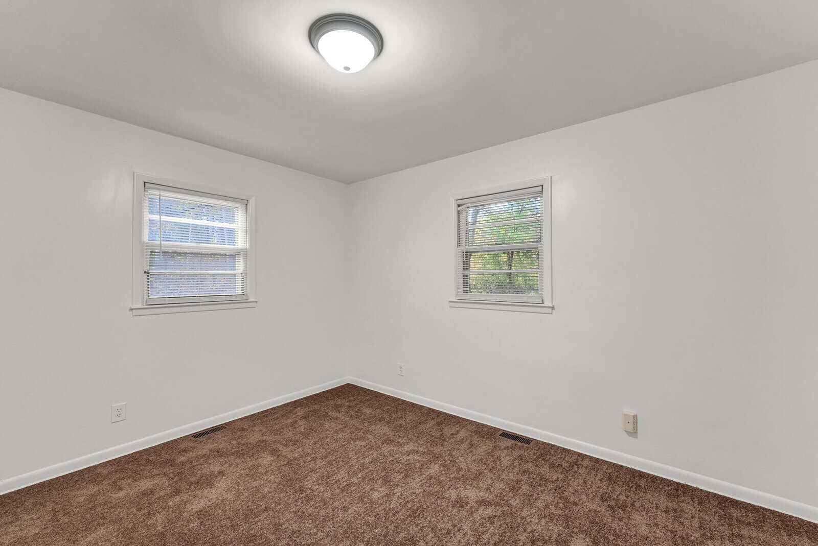 An empty room with white walls, two windows with blinds, and brown carpet flooring.