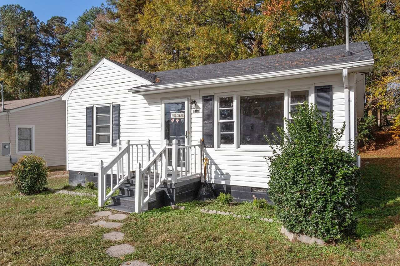 A small, white single-story house with black shutters, featuring a set of stairs leading to the front door and a green lawn with a bush to the side.