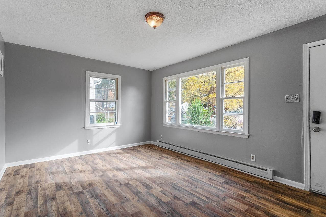 Empty room with gray walls, wooden flooring, and a view of trees with yellow foliage through a large window.