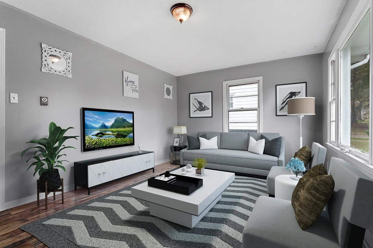A well-appointed living room featuring a grey sofa, matching armchair, a white coffee table, and entertainment unit with a large television, complemented by grey walls adorned with various picture frames, and a zigzag-patterned area rug. light streams in from a window to the left, and the room has a ceiling-mounted light fixture.