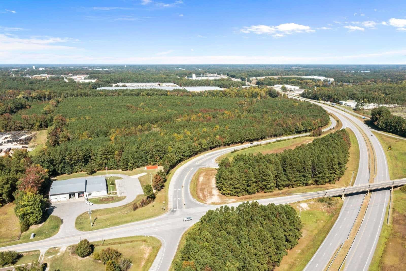 Aerial view of a curving road with an overpass near a rural landscape with mixed patches of trees and industrial buildings.