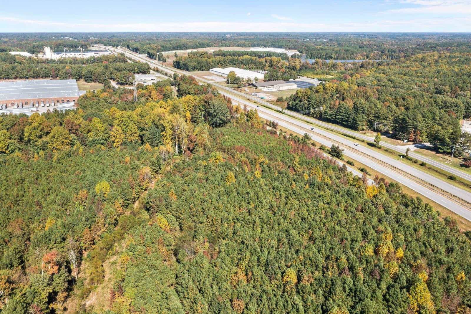 Aerial view of a highway running through a forested area with early autumn foliage and industrial buildings in the distance.