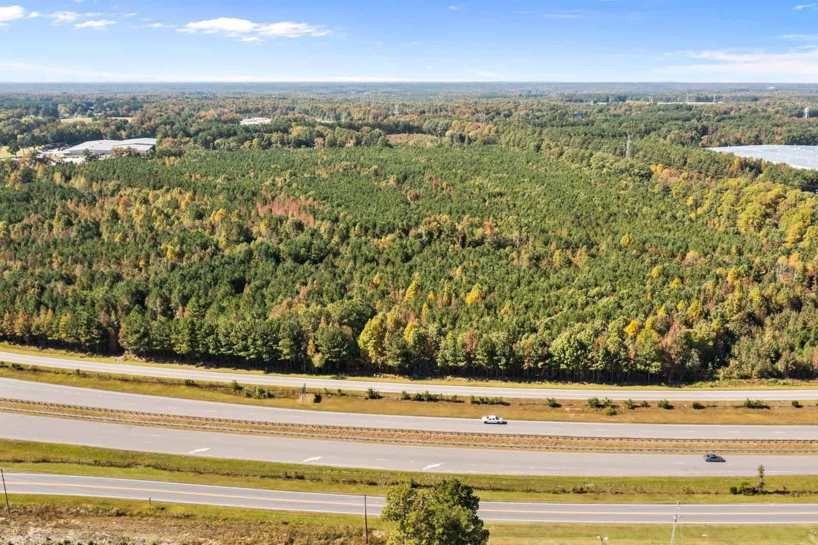 Aerial view of a highway running parallel to a vast forest with trees showing early autumn colors, under a clear blue sky.