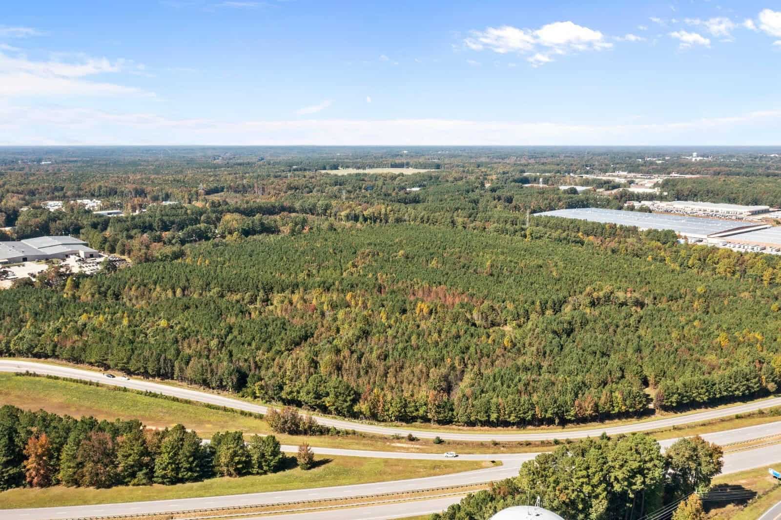 Aerial view of a highway junction with surrounding forested area and industrial buildings in the distance.