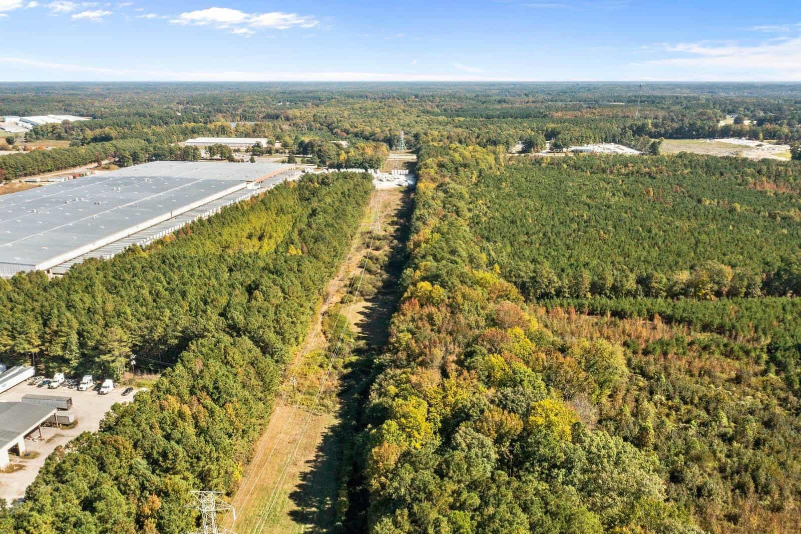 Aerial view of a tree-lined path dividing two large areas, with industrial buildings on the left and a dense forest on the right under a clear sky.
