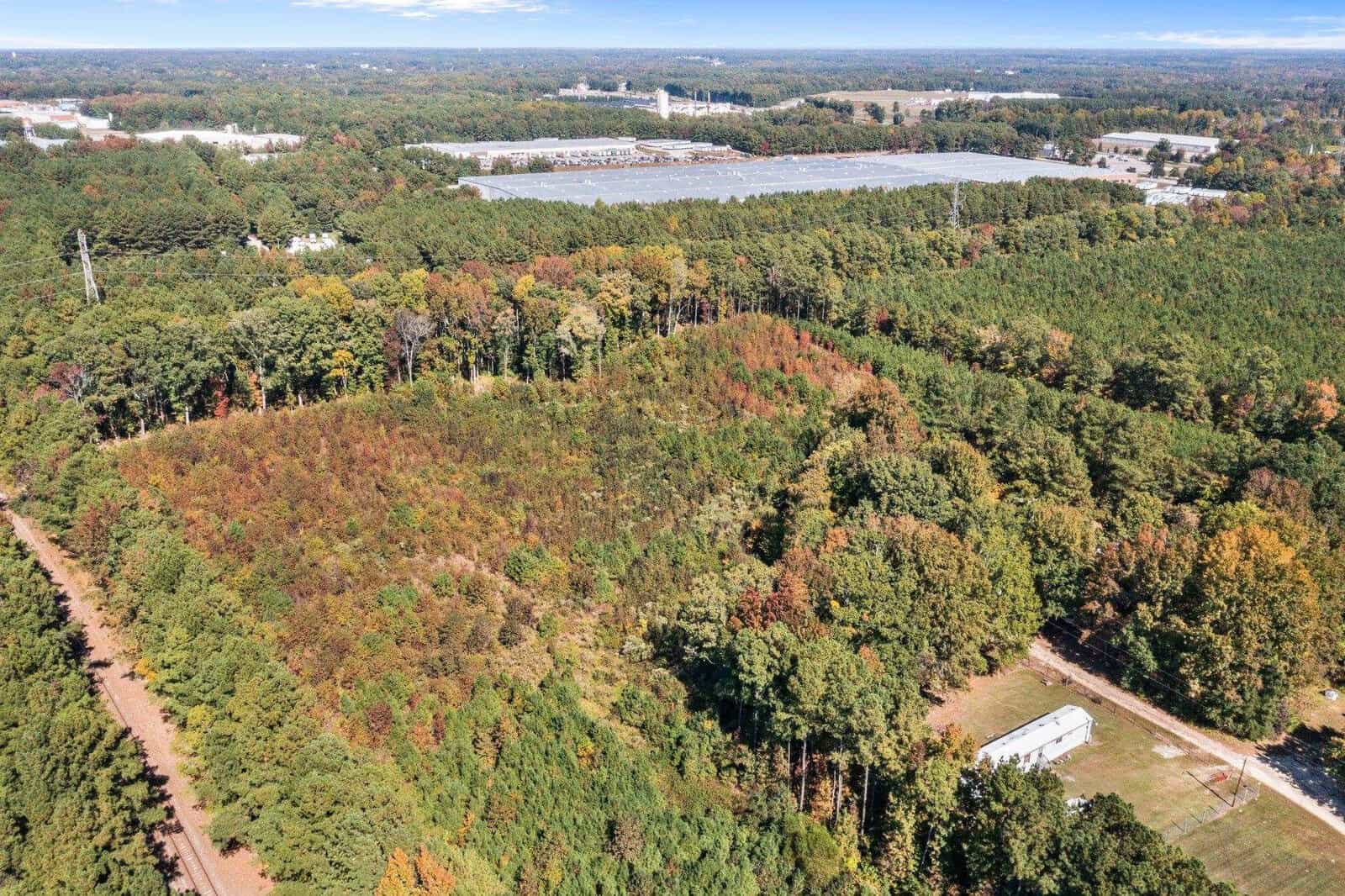 Aerial view of a mixed forest with early autumn colors adjacent to a railway line, with industrial buildings in the background.