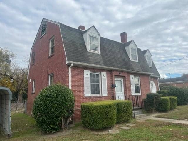 A red brick, two-story house with white-trimmed windows and a shingled roof, featuring dormer windows and surrounded by shrubbery, under a cloudy sky.
