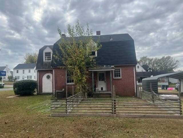A two-story house with a brick facade on the lower level and white siding on the upper level, featuring a front entrance partially covered by an overgrown tree, with a weathered wooden fence in the foreground and a carport to the side, all under a cloudy sky.