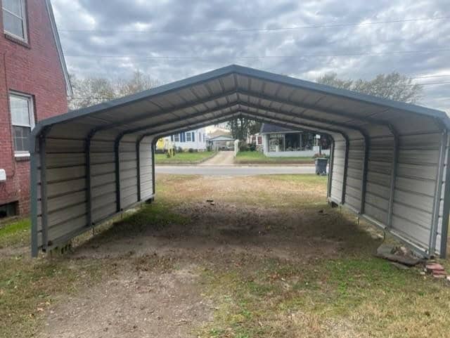 A metal carport with no vehicle parked under it, situated on a grassy area with residential structures in the background.