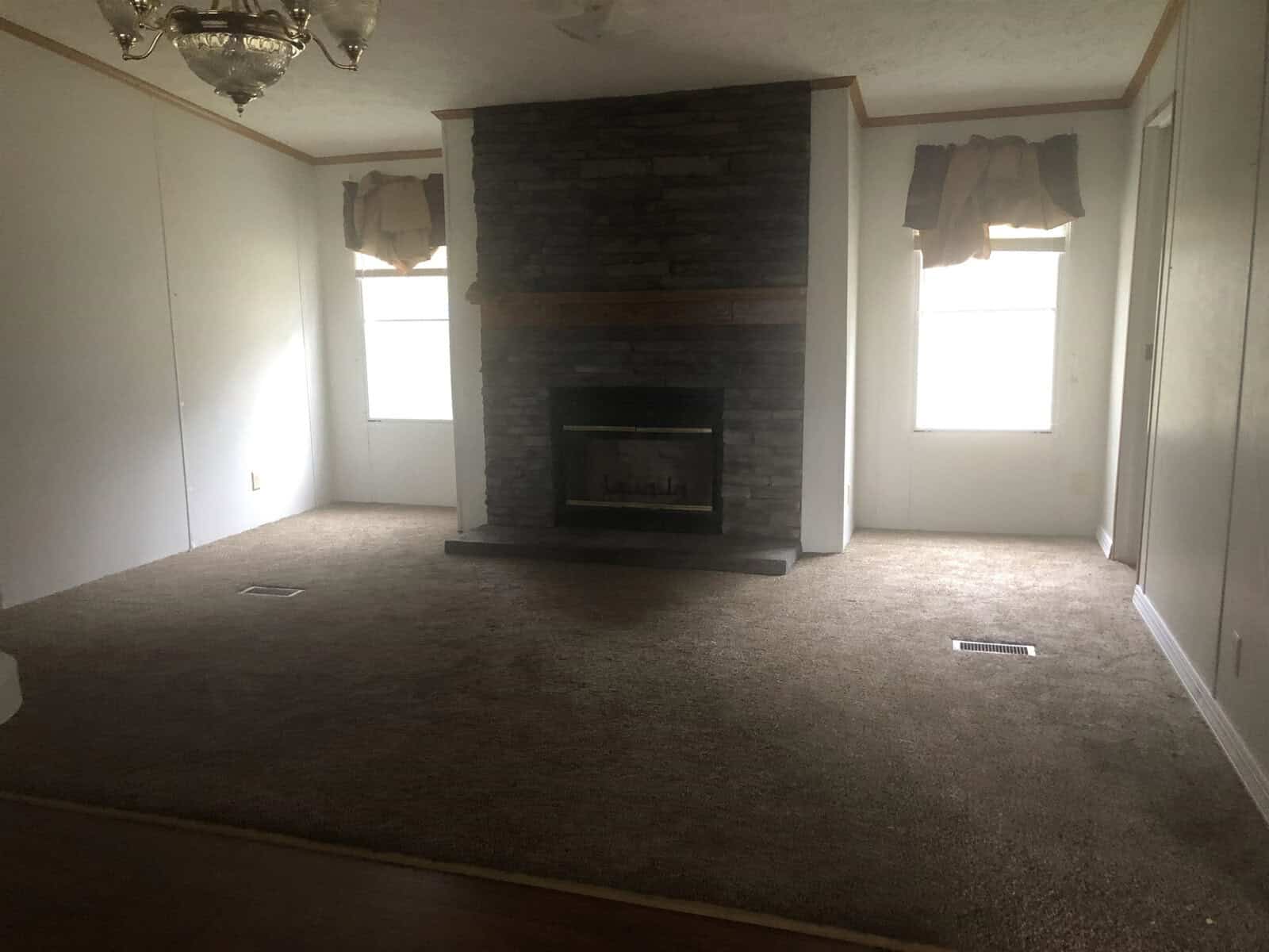 An empty room with beige carpeting featuring a central brick fireplace and two windows with valances.
