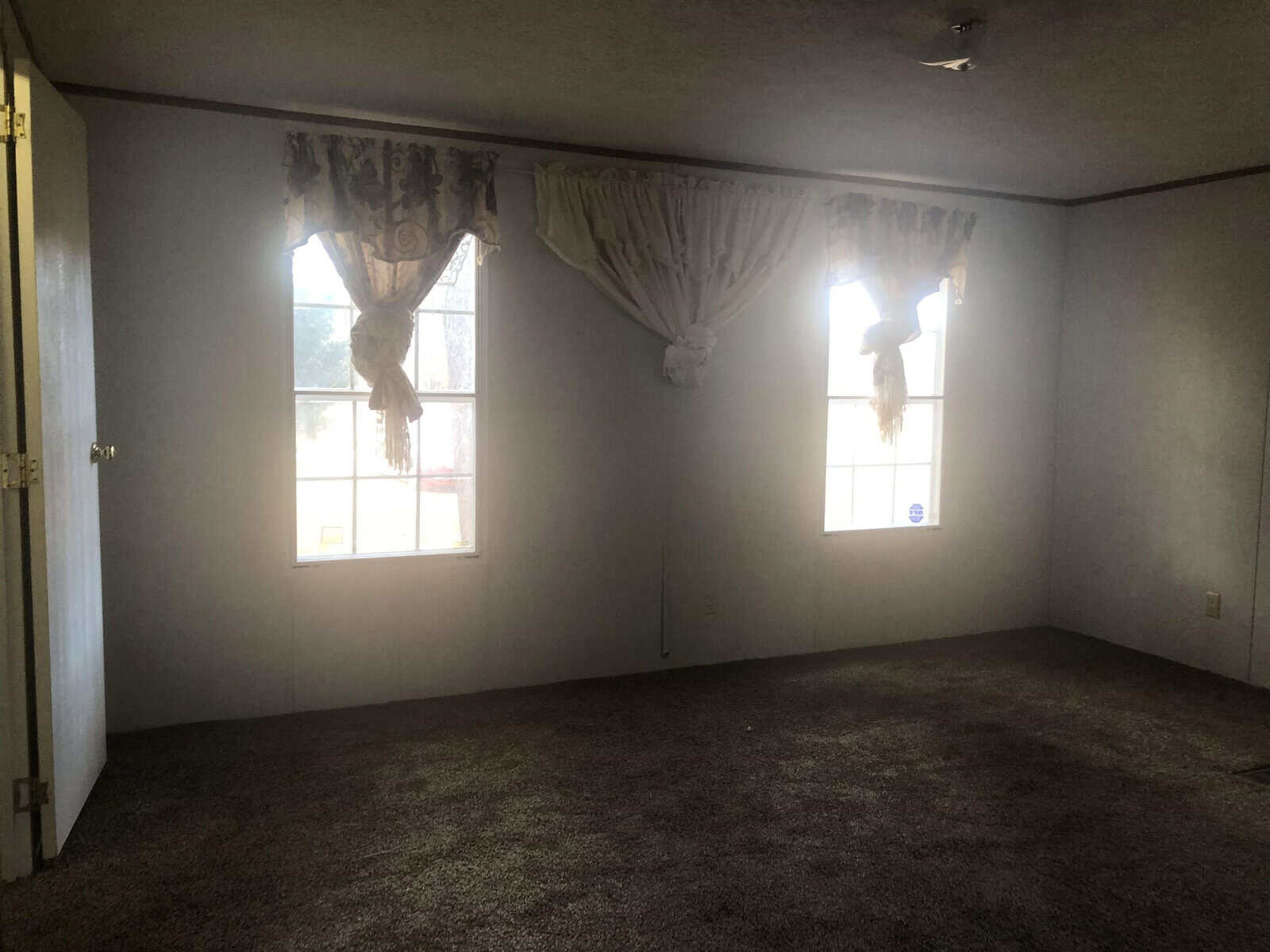 An empty room with carpeted floor, two windows with draped curtains, and visible sunlight coming through.