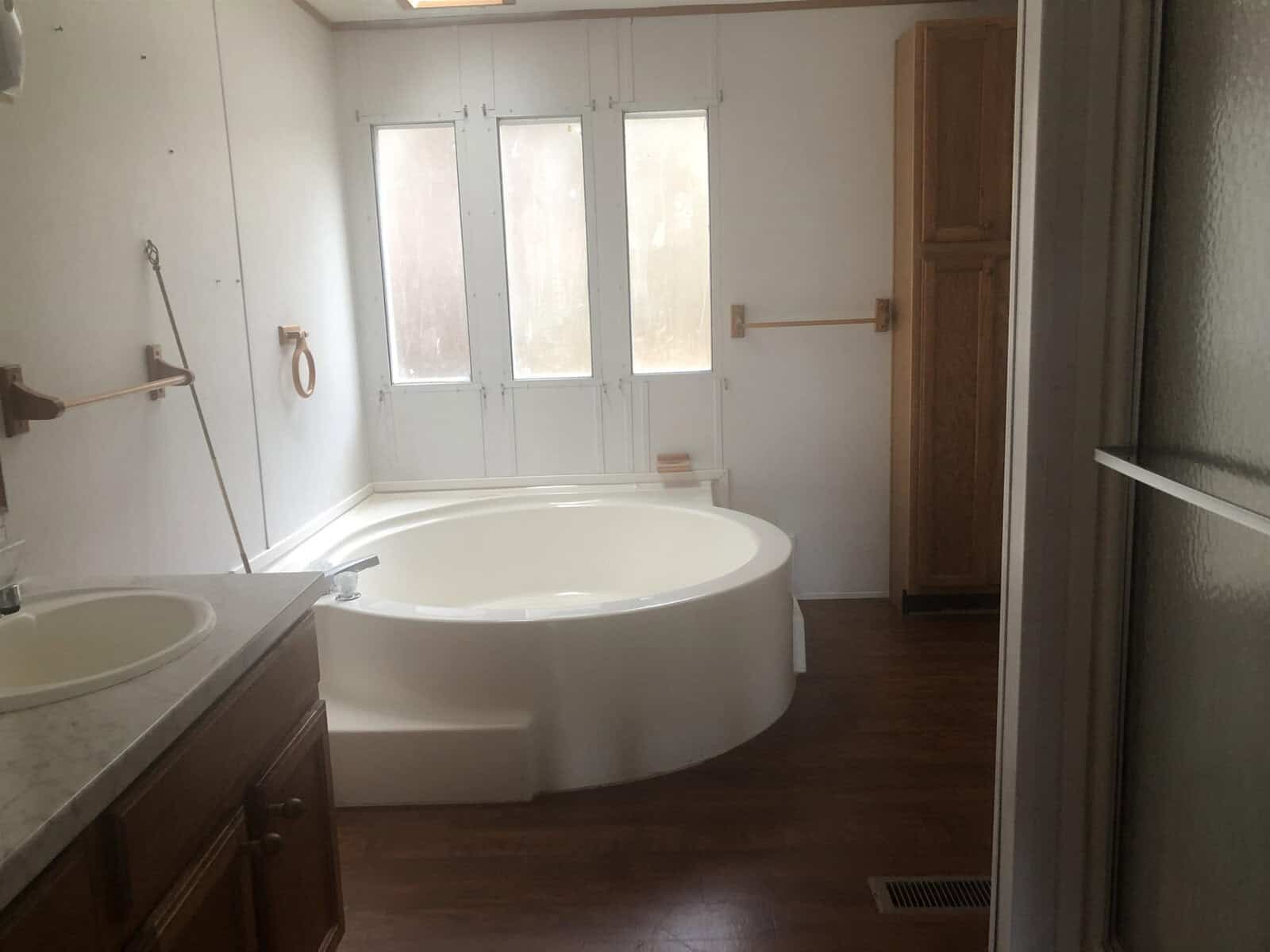 A spacious bathroom featuring a large corner bathtub, a double sink vanity with a marble countertop, frosted glass windows for privacy, and wooden cabinetry.