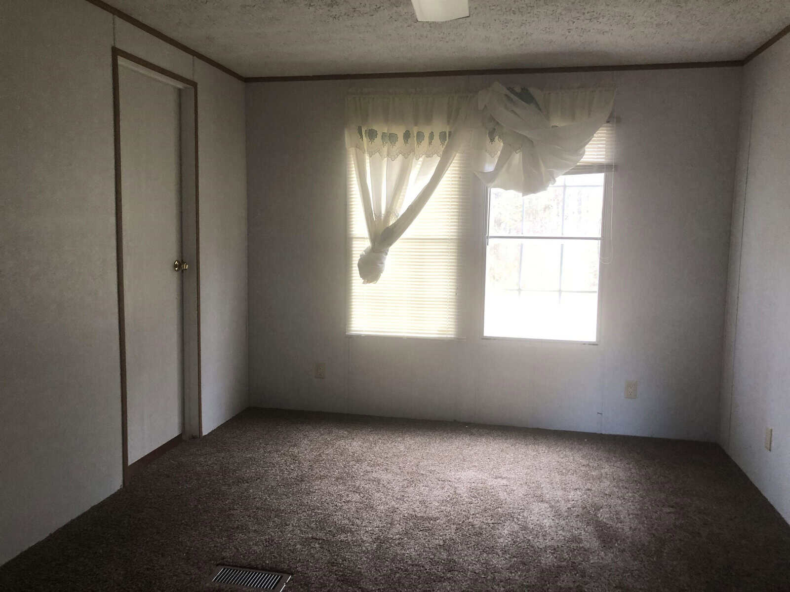 A vacant room with a single window covered by a partially fallen curtain, beige carpeting, and a closed door on the left wall.