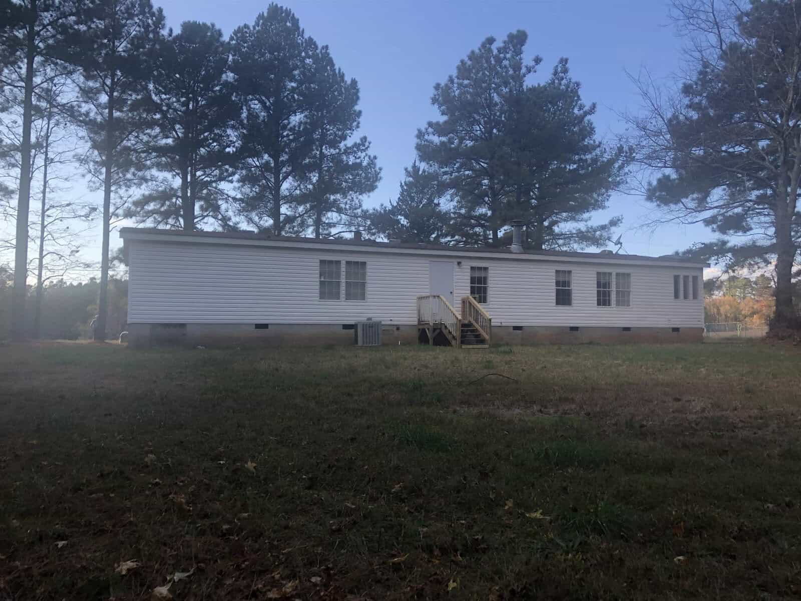 A single-story white mobile home with wooden steps on a grassy area surrounded by trees.