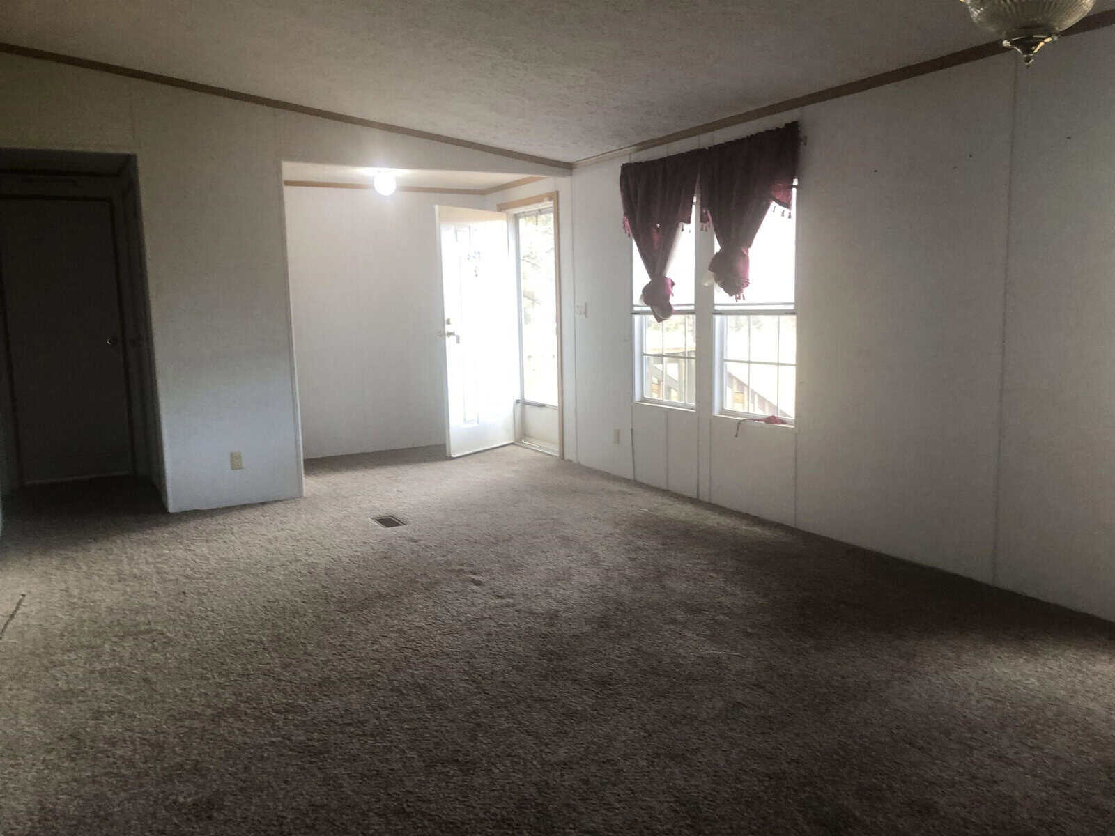 An empty room with beige carpeting featuring a glass sliding door leading to the outside, adorned with maroon curtains, and an overhead ceiling fan.