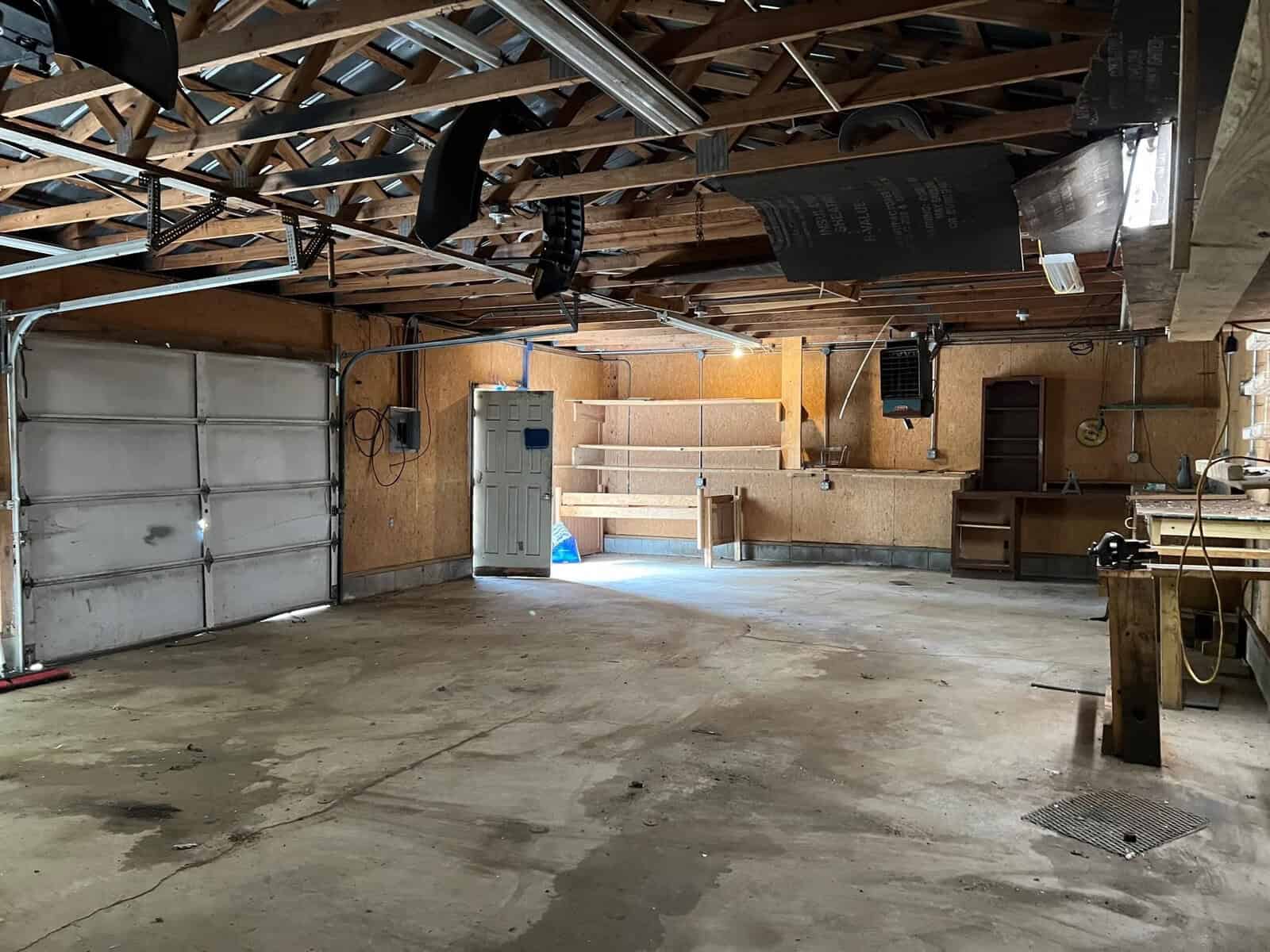 An empty garage with an open door, featuring exposed wooden rafters, storage shelves along the back wall, and a concrete floor with visible stains.