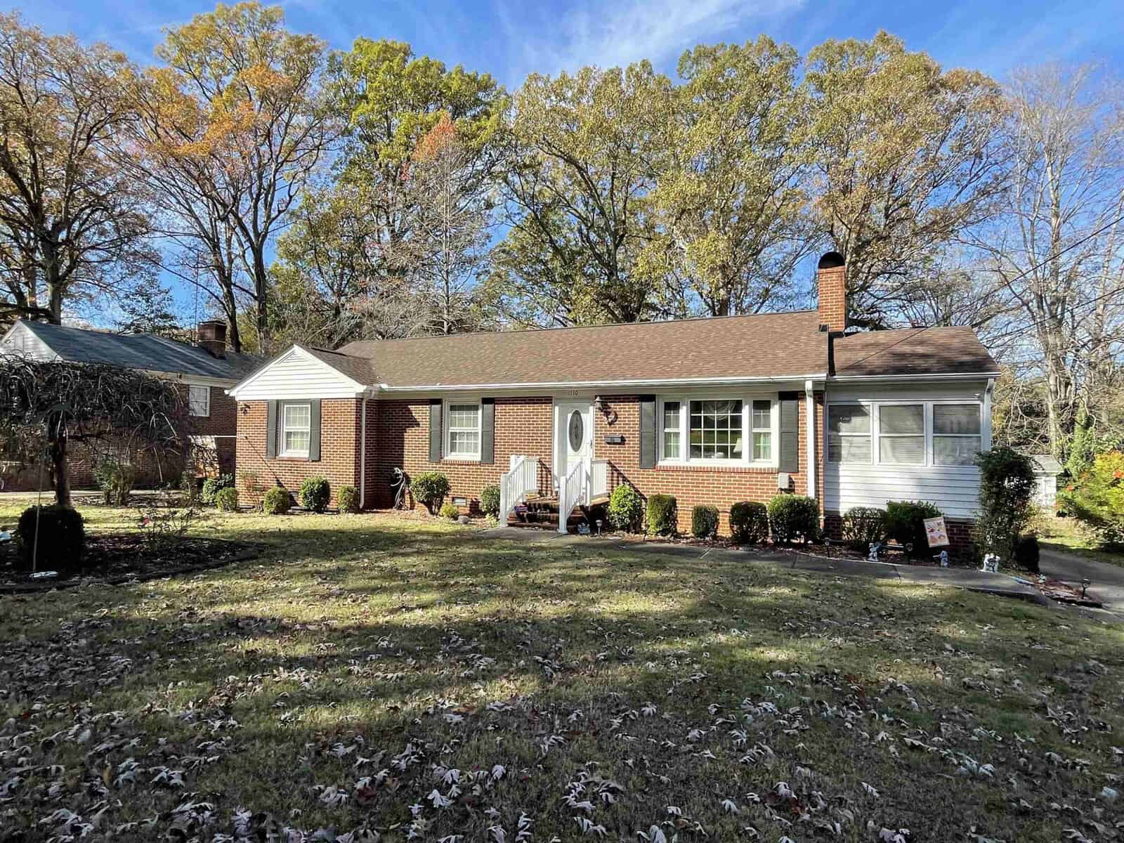 A single-story brick house at 1510 Oakdale Circle, Henderson NC, with white trim, featuring a central white door with side panels, flanked by two large windows, and a chimney on
