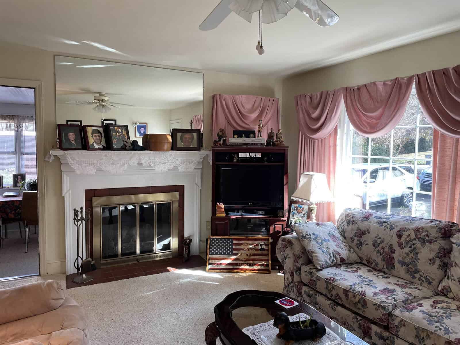 A traditional living room with a floral-patterned couch, a glass coffee table, and a fireplace with framed photos on the mantelpiece. the room features draping pink curtains, a ceiling fan, and a television set within a wooden cabinet. bright daylight filters through the windows.