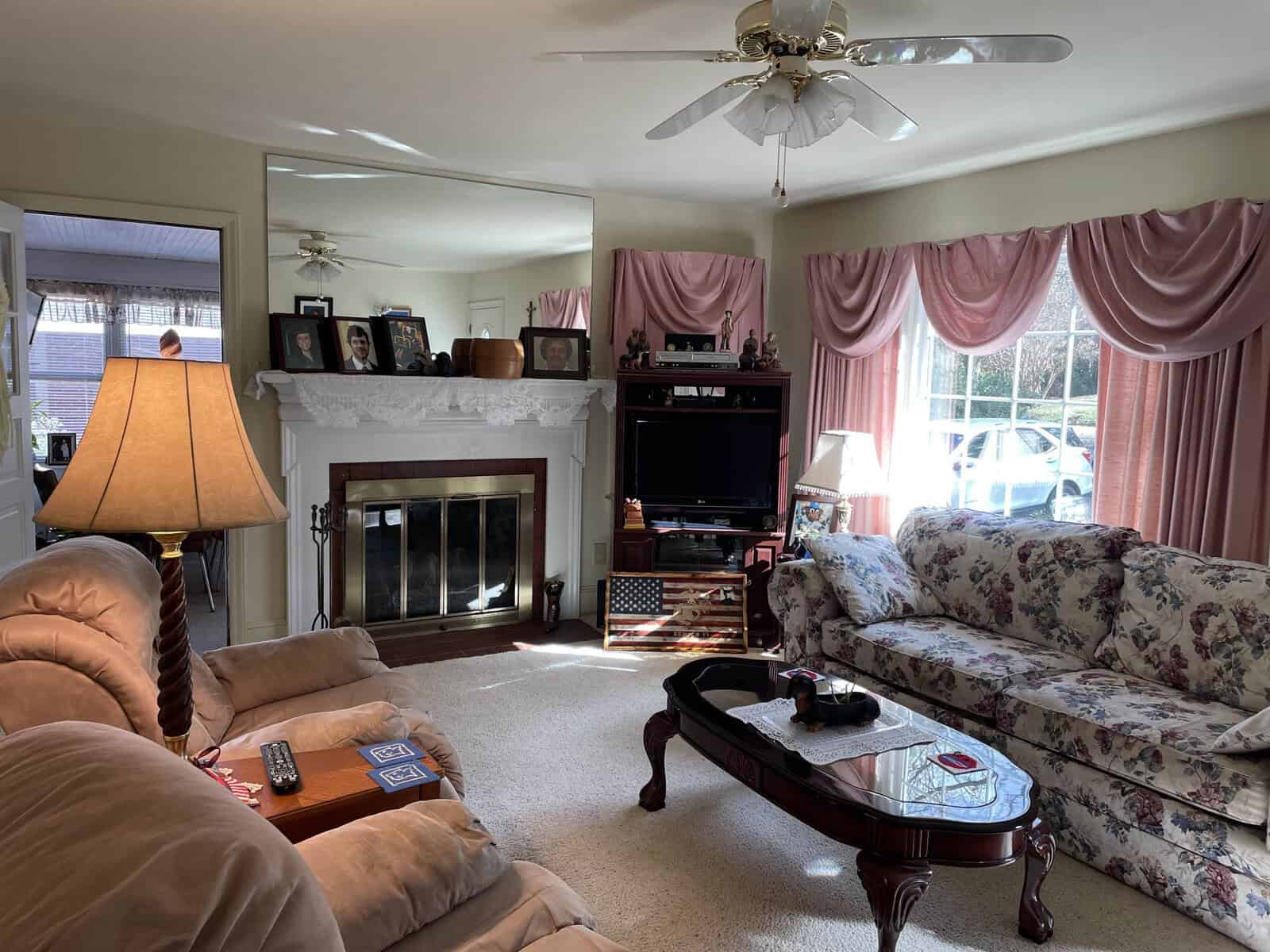 A cozy living room featuring a fireplace, a sofa with a floral pattern, a loveseat, a wooden coffee table, a television set on a black stand, and draped pink curtains on the windows.