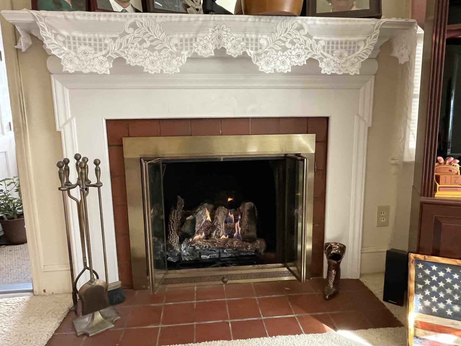 A traditional fireplace with a white mantel and gold fireplace screen, accompanied by a set of fireplace tools on the left side, is situated in a living room setting with decorative items on the mantel and an american flag partially visible on the right.