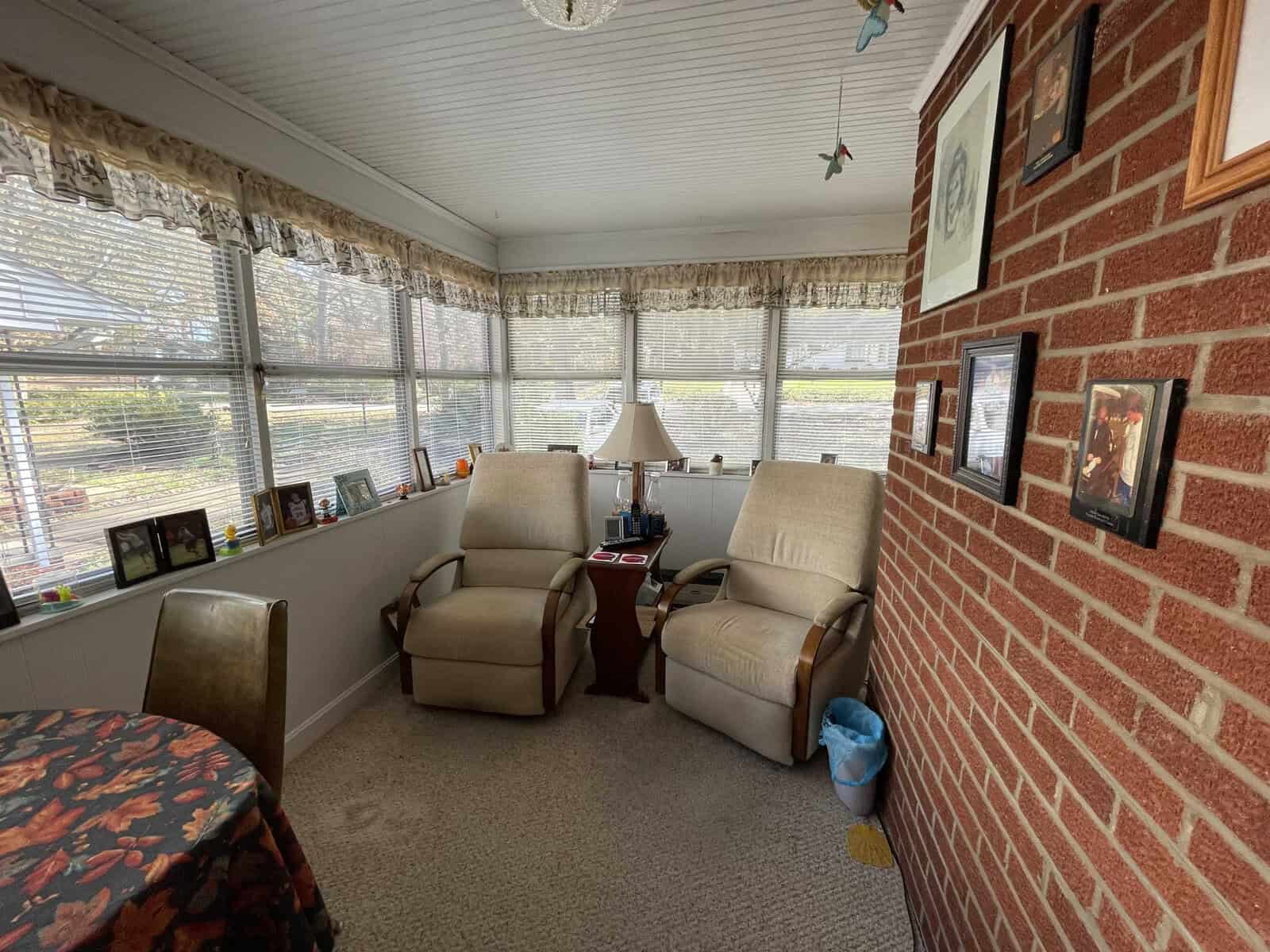 A cozy sunroom with two recliner chairs, brick wall on one side, lace curtains on the windows, and various decorations and photographs.