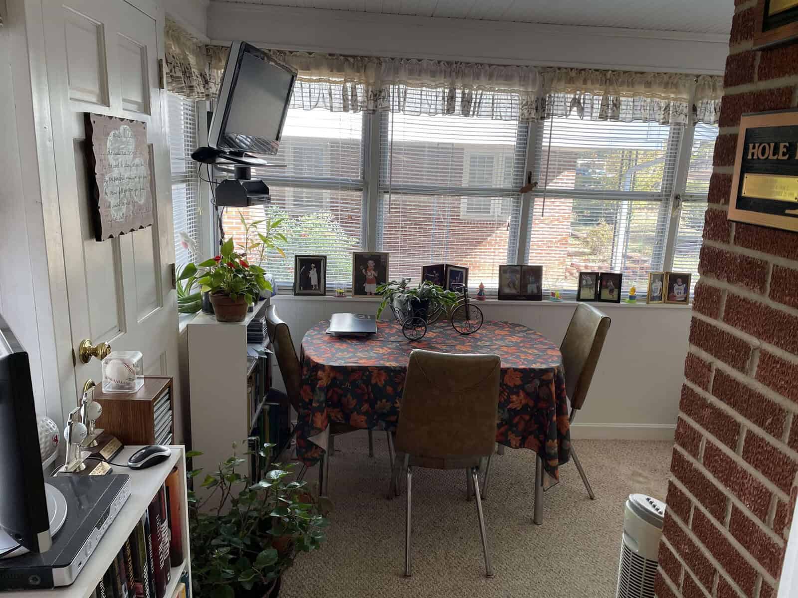 A cozy home office setup with a desk and computer by the window, surrounded by personal photographs and decorative items, with a table and chairs at the center of the room, featuring brick walls and a view of the exterior environment through the windows.