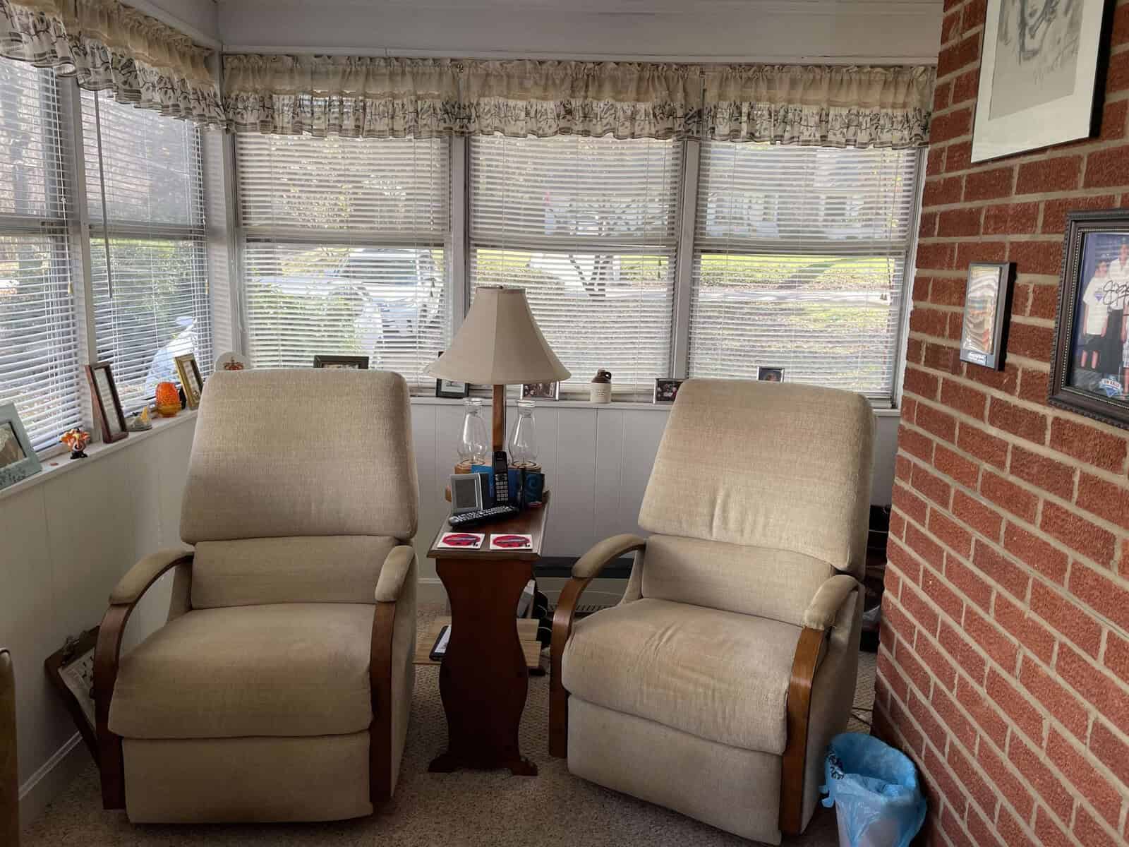 A cozy sitting area with two upholstered chairs, a side table with a lamp and books, situated near a weathered brick wall adjacent to a window with blinds and patterned curtains.