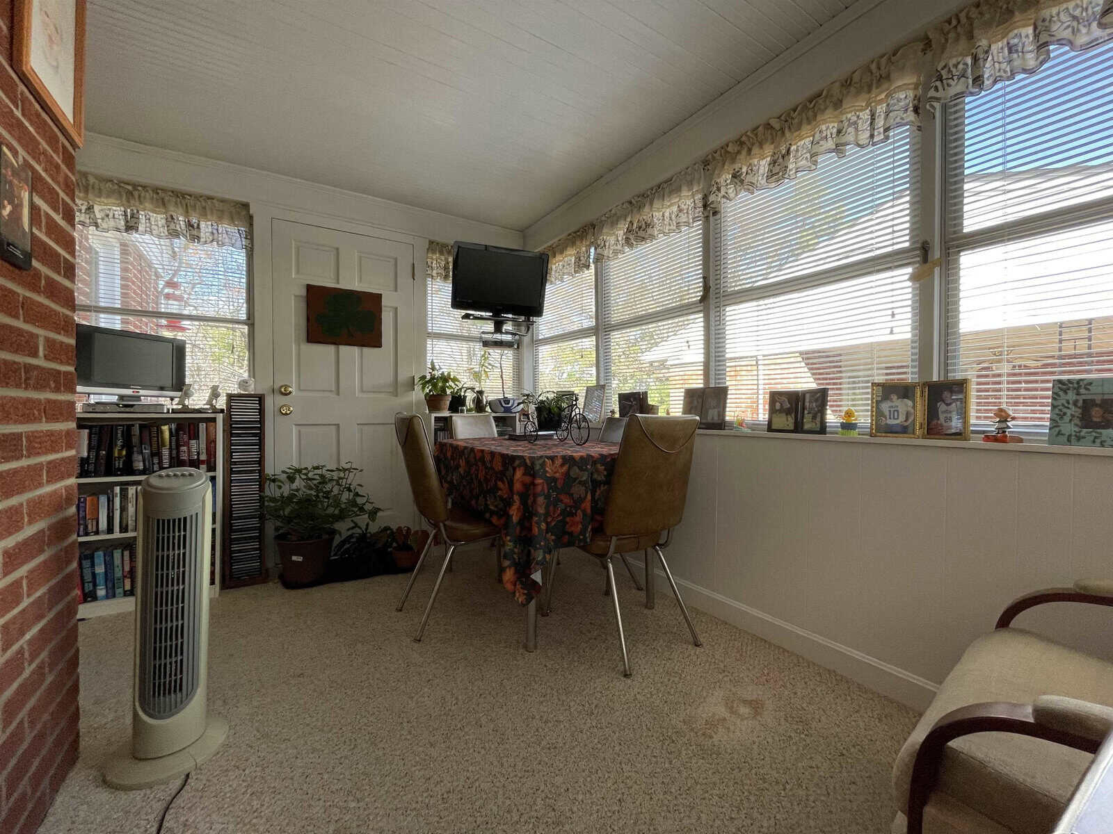 A well-lit, cozy room with a dining table and chairs, a tv mounted on the wall, bookshelves, plants, and an oscillating fan.