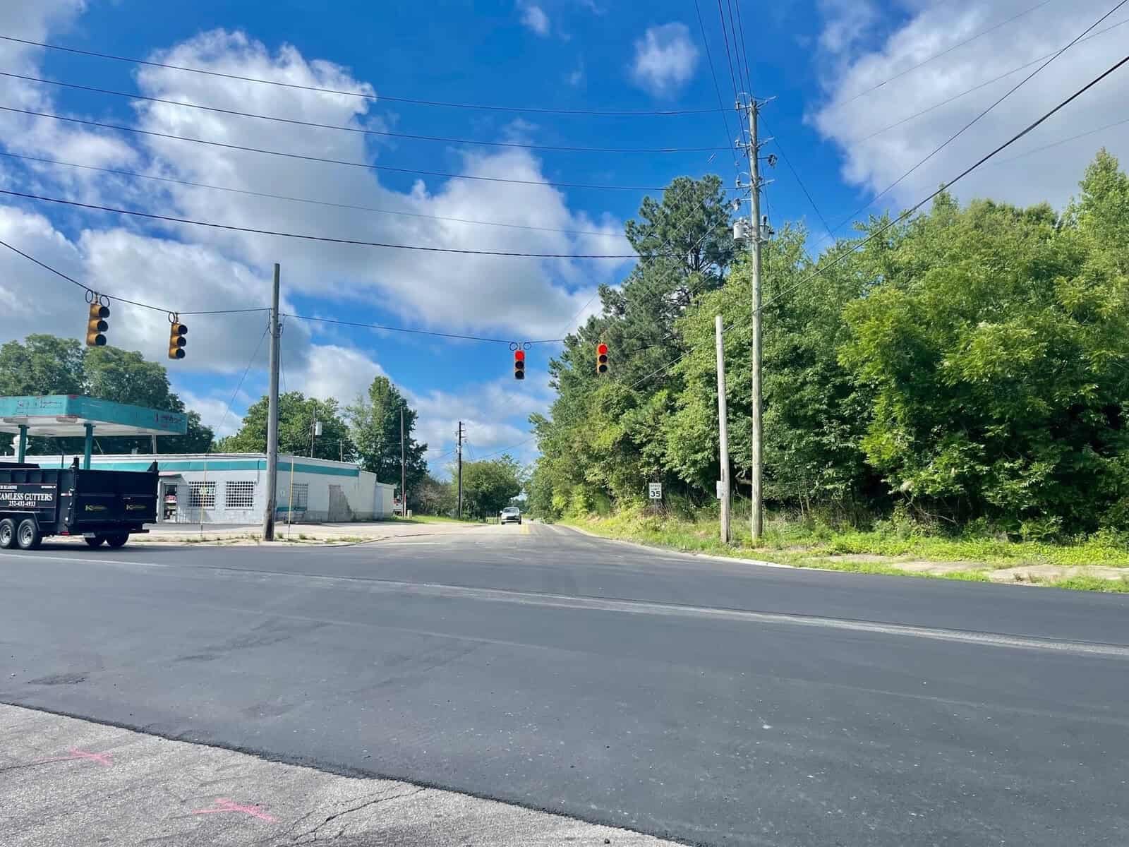 Intersection with red traffic lights and a white truck turning, surrounded by lush greenery and a clear blue sky. a gas station is visible on the left.