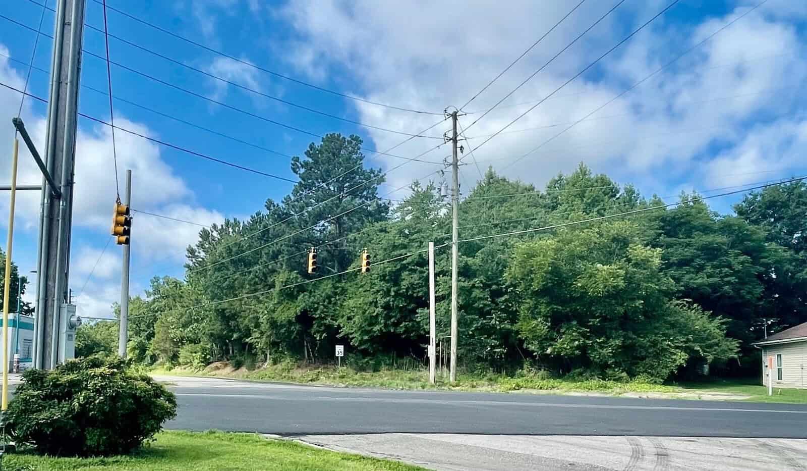 A suburban intersection with hanging traffic lights and a lush tree background under a partly cloudy sky. electrical poles line the street, and there is a small building on the right.