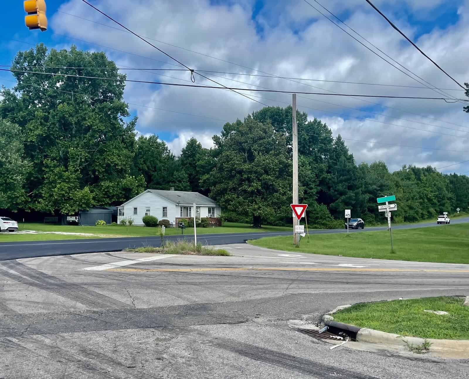 A suburban intersection featuring a yield sign, wide streets, and a white house surrounded by greenery under a cloudy sky. traffic lights and utility poles are also visible.