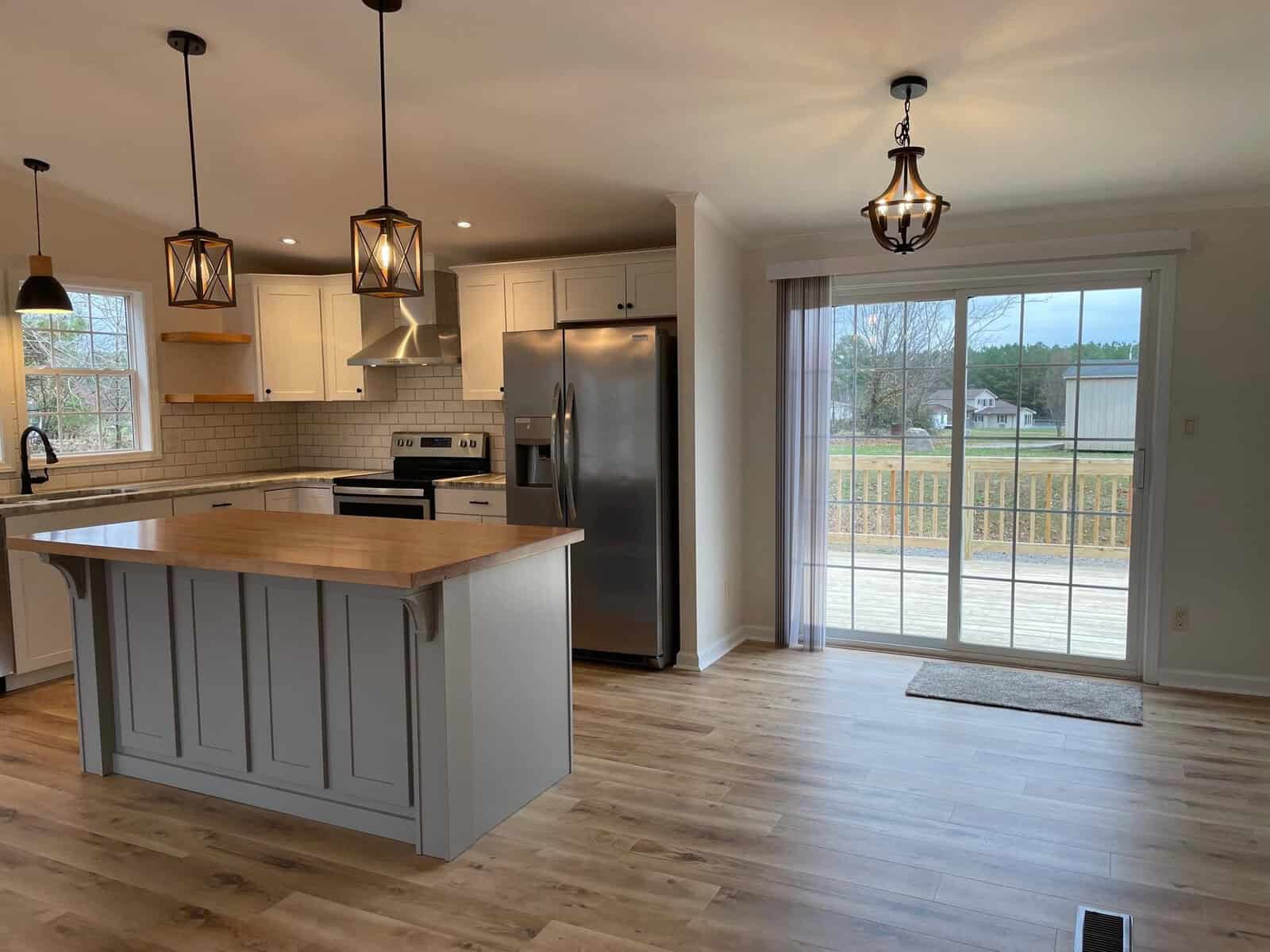A modern kitchen with stainless steel appliances, white cabinetry, and an island with pendant lighting, opening onto a room with a chandelier and a sliding glass door leading outside.