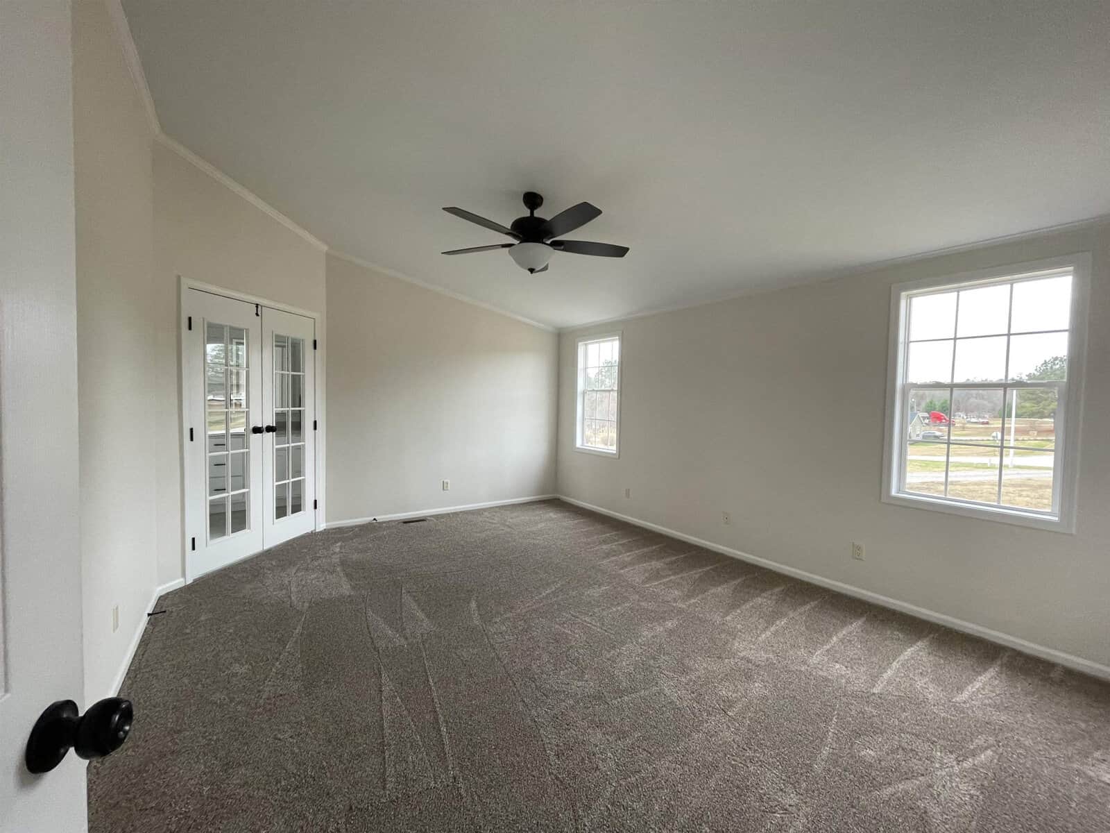 An empty room with beige carpeting, white walls, a ceiling fan, two windows providing natural light, and a closed white door with glass panes.