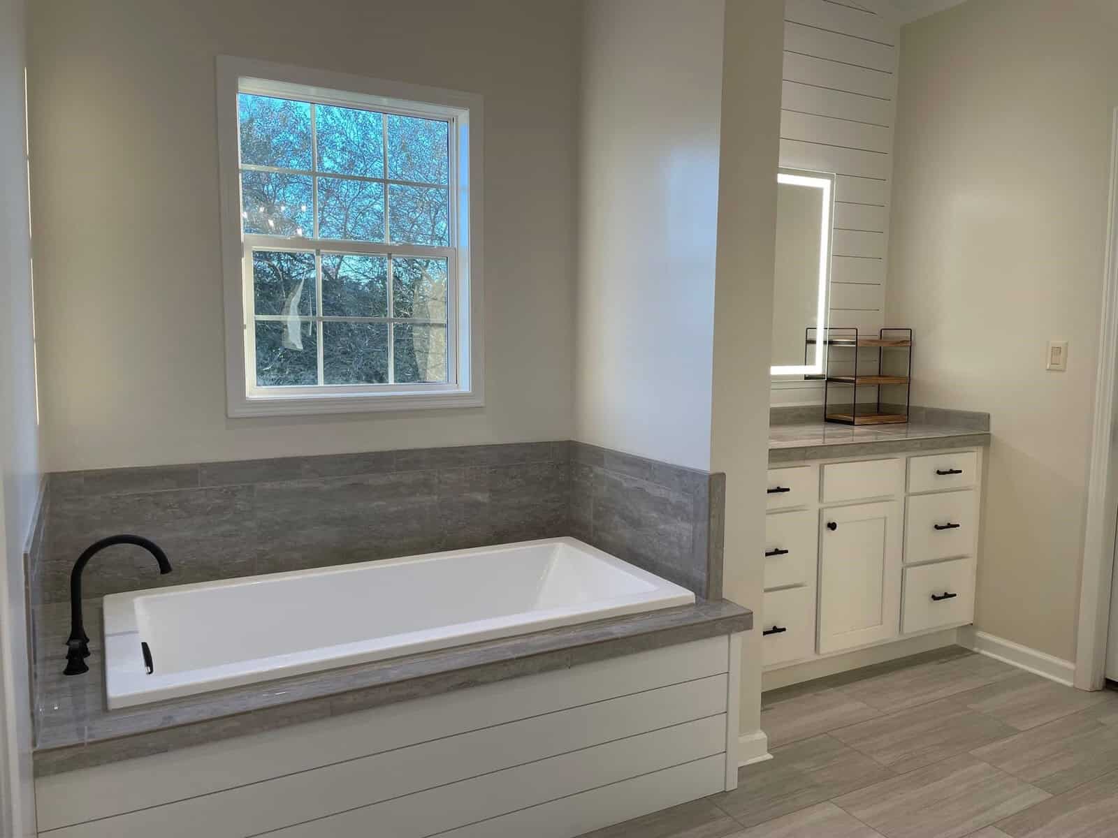 Modern bathroom with a built-in bathtub, gray tiling, white cabinetry, and a window allowing natural light.