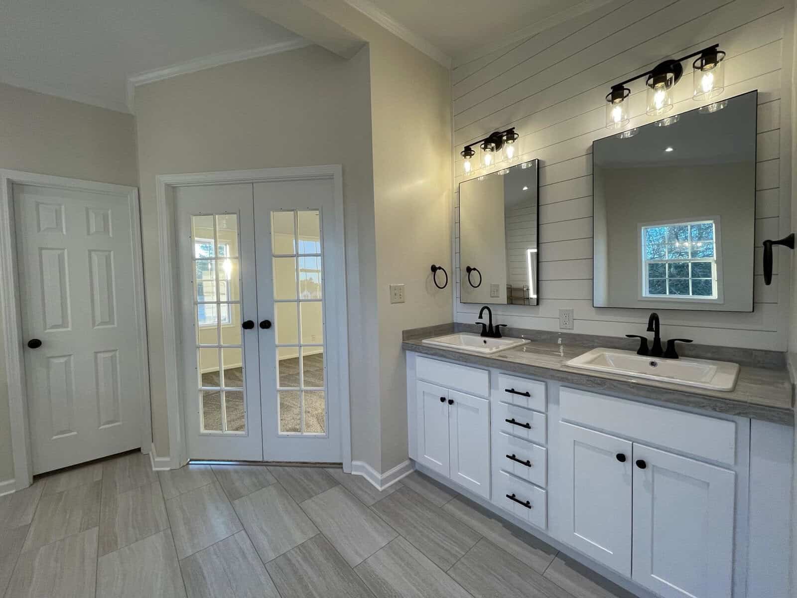 A modern bathroom interior with double sinks, white cabinetry, large mirrors with overhead lighting, and gray flooring. the room features a frosted glass door and a window providing natural light.