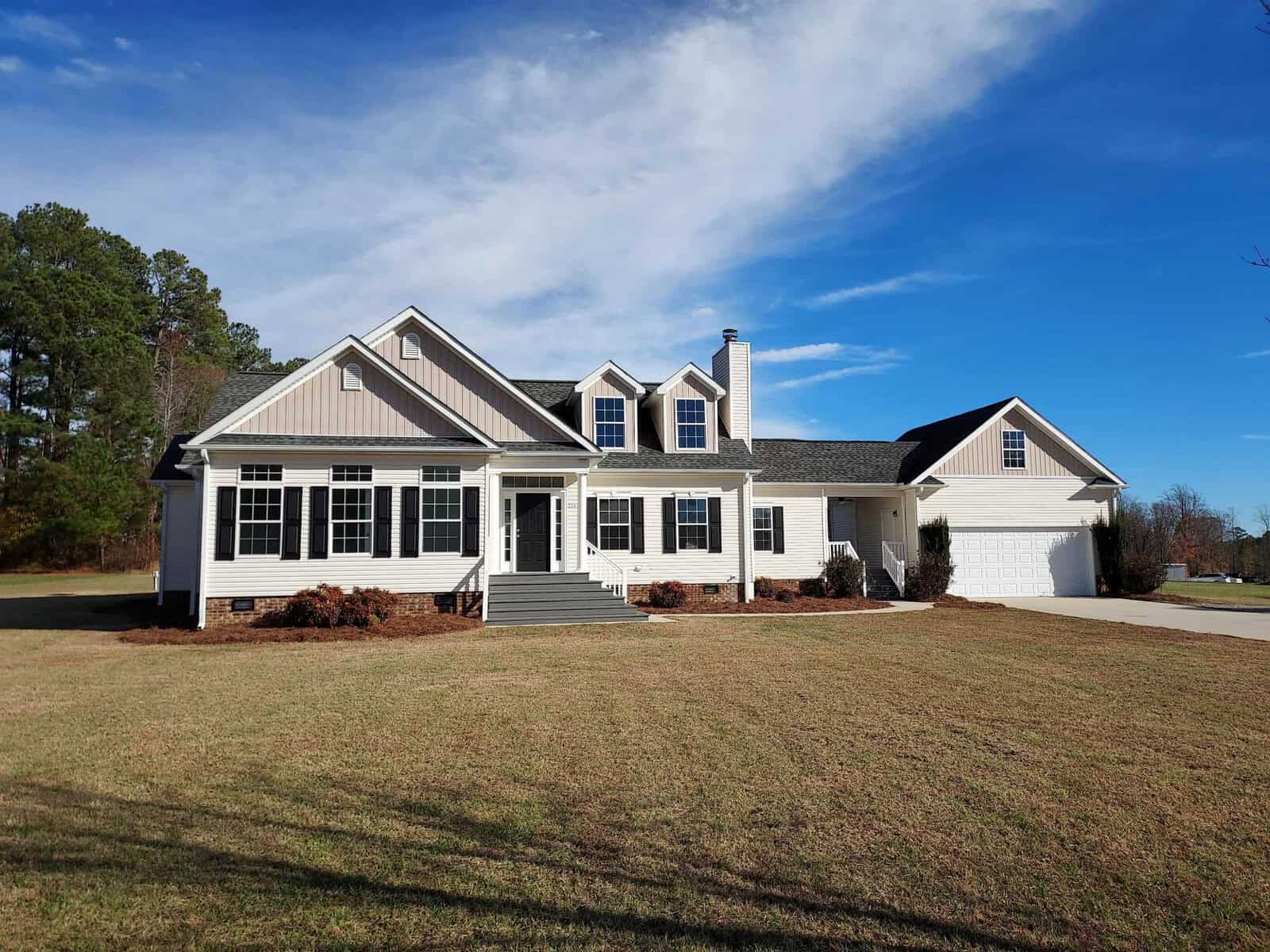 A spacious, modern suburban home at 2243 Bobbitt Road in Kittrell with white siding, black shutters, and a large front lawn under a clear blue sky. The house features a two-story design with multiple gables, large windows, a chimney, and an attached two-car garage. Trees are visible in the distance.