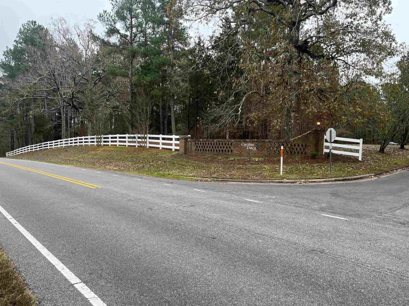 A quiet, tree-lined street corner with a white wooden fence following the curve of the road, a mailbox, and some residential signage.