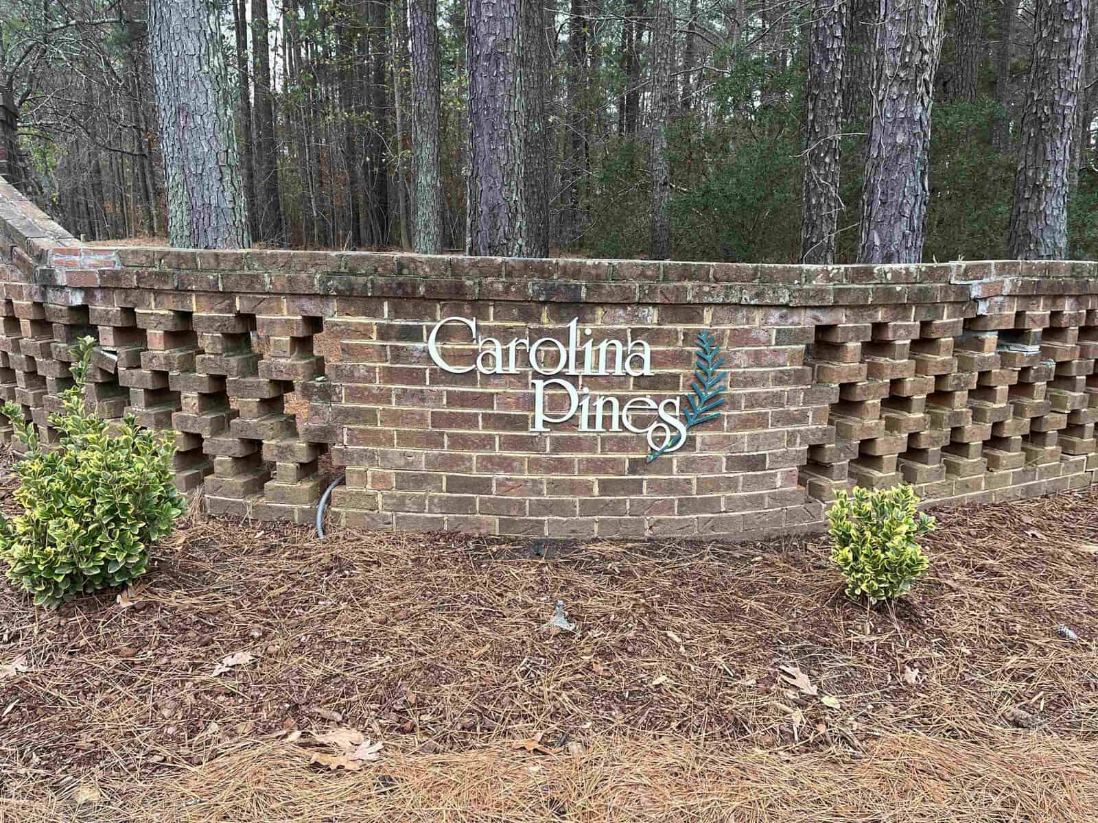 Brick wall with the words "carolina pines" written on it, flanked by shrubbery and surrounded by pine trees.