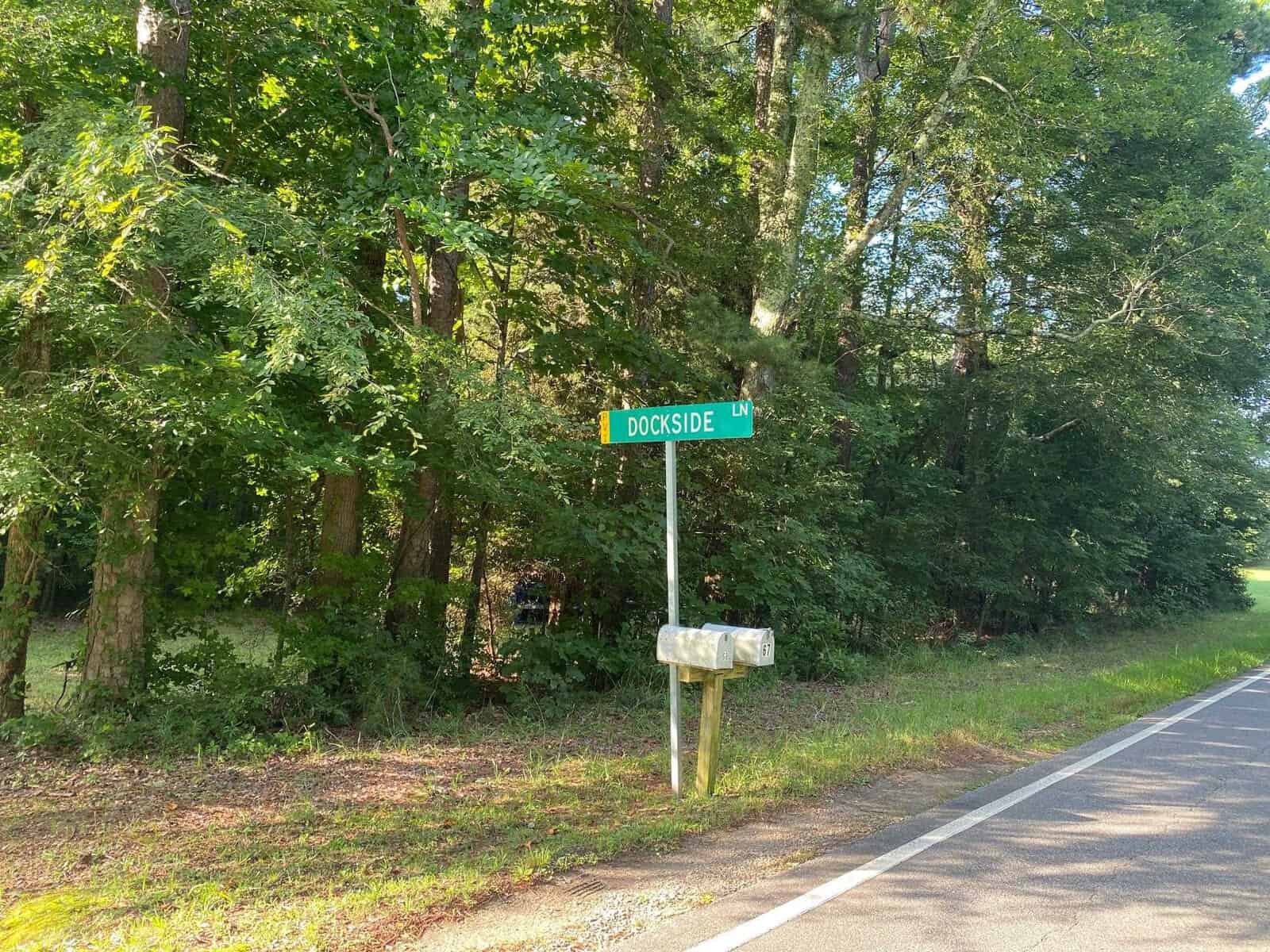 A street corner with a green street sign that reads "dockside ln" next to a rural road with mailbox and lush green trees in the background.