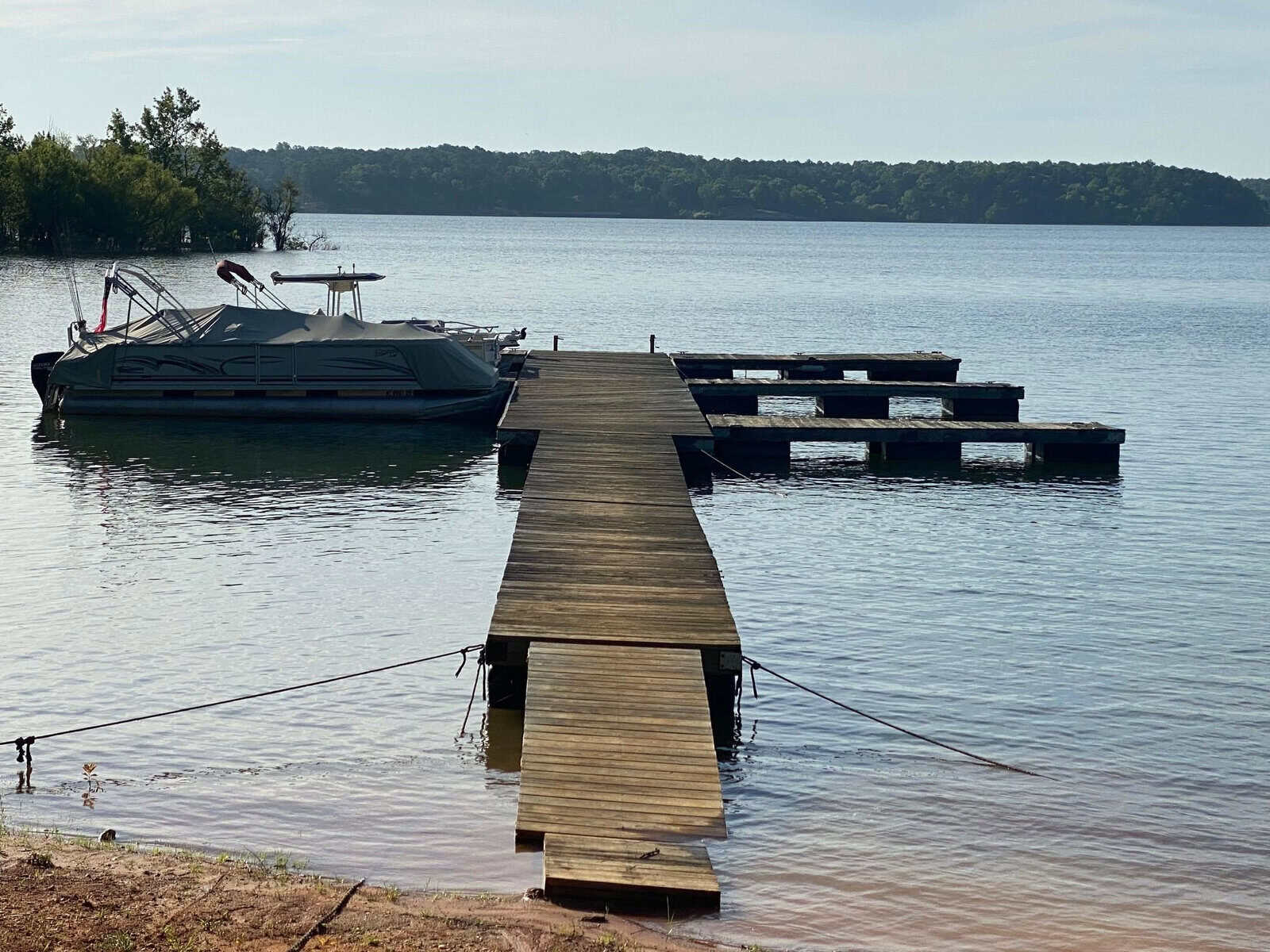 A pontoon boat moored to a wooden dock on a calm body of water with surrounding trees.