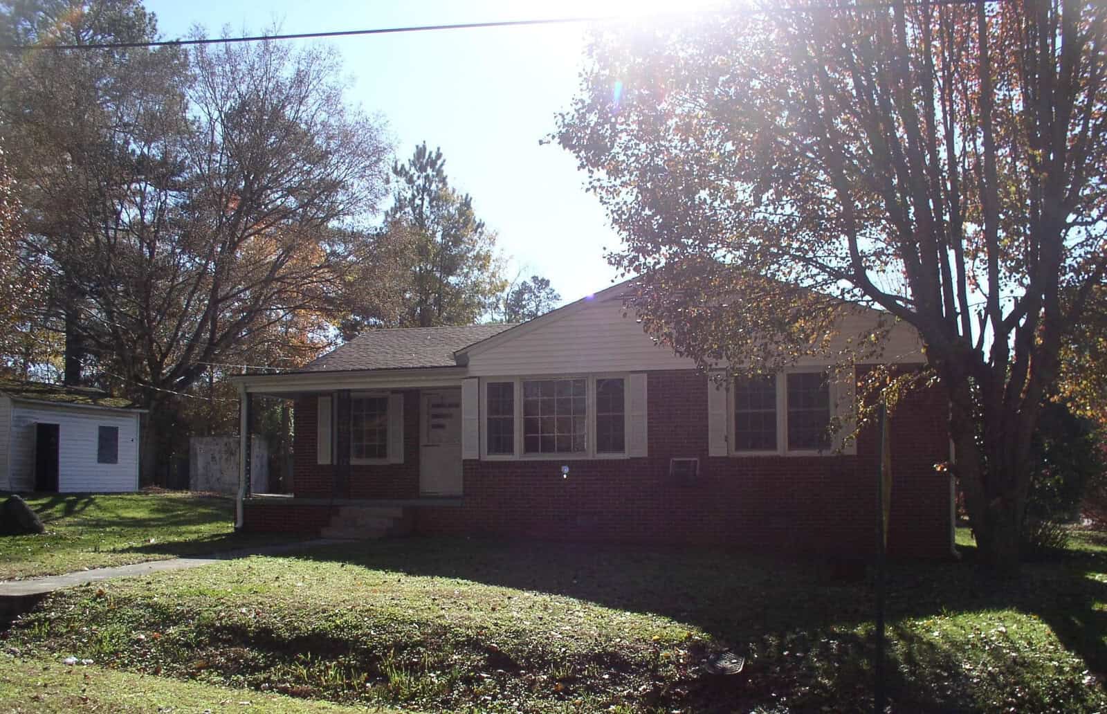A single-story red brick house with white trim and a small porch under a clear blue sky, surrounded by green grass and trees with autumn foliage.