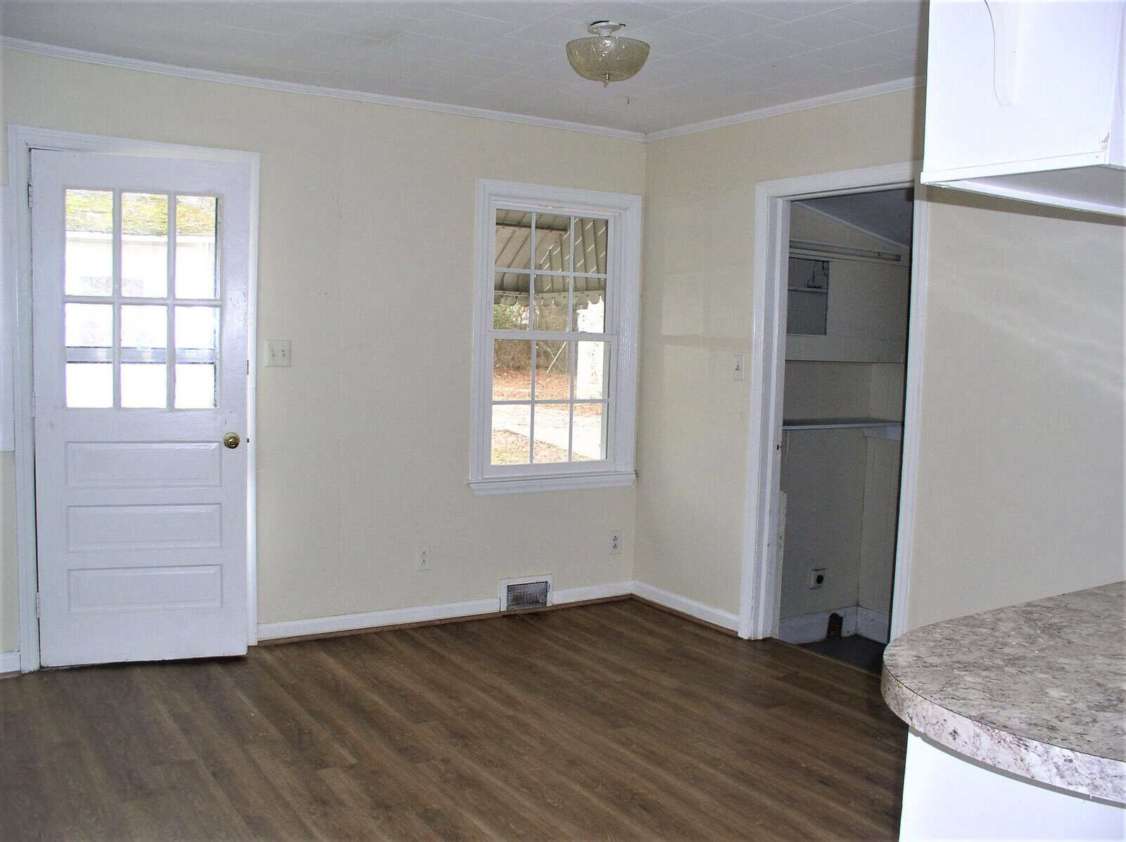 An empty room featuring a wood-like laminate floor, a white door with a window, a closed closet, and a section of a kitchen counter on the right side.