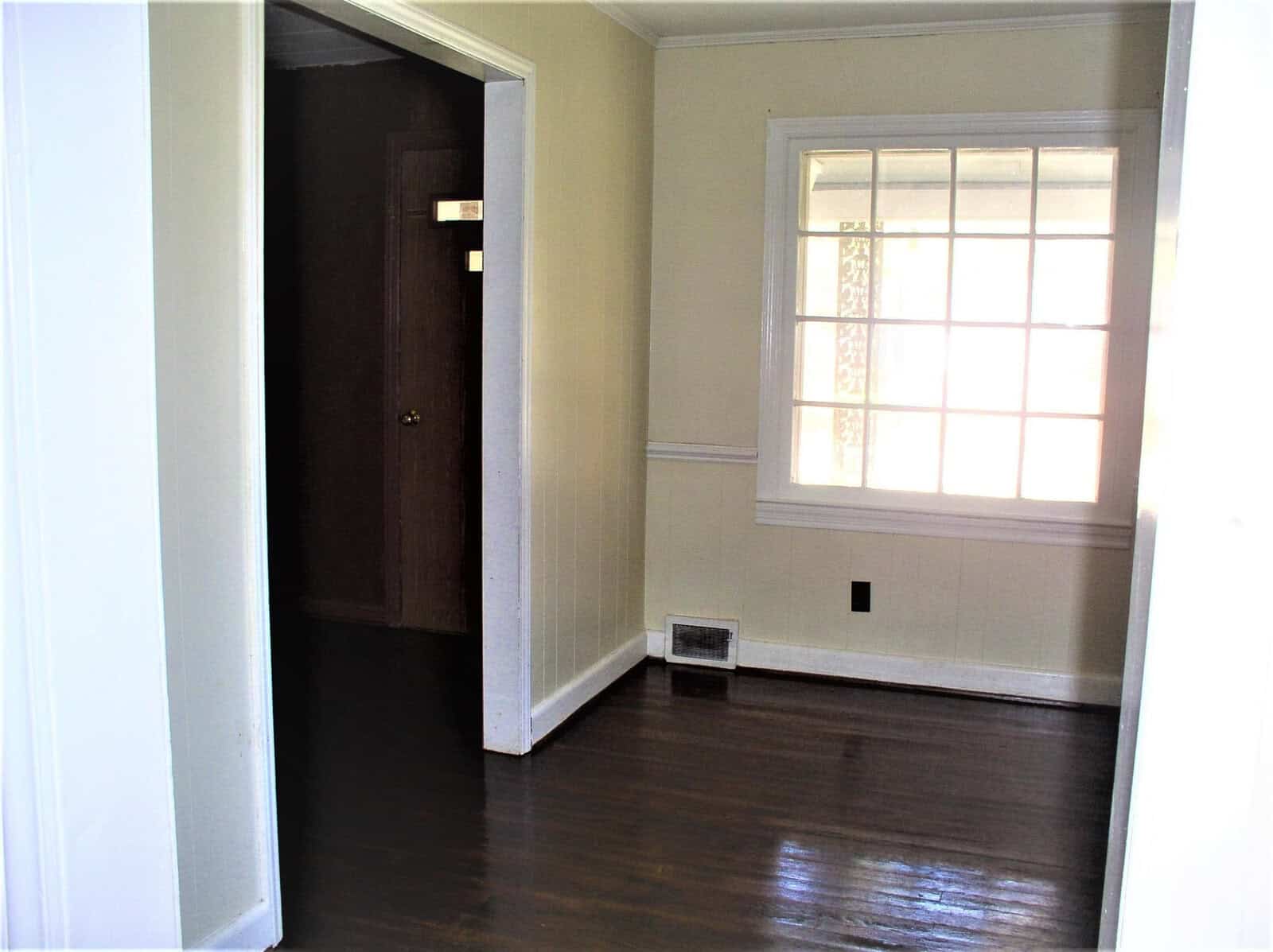 An empty room with polished wooden floors, white walls, and a single window with translucent curtains, casting soft light into the space. an open doorway leads to another room with a closed brown door visible.
