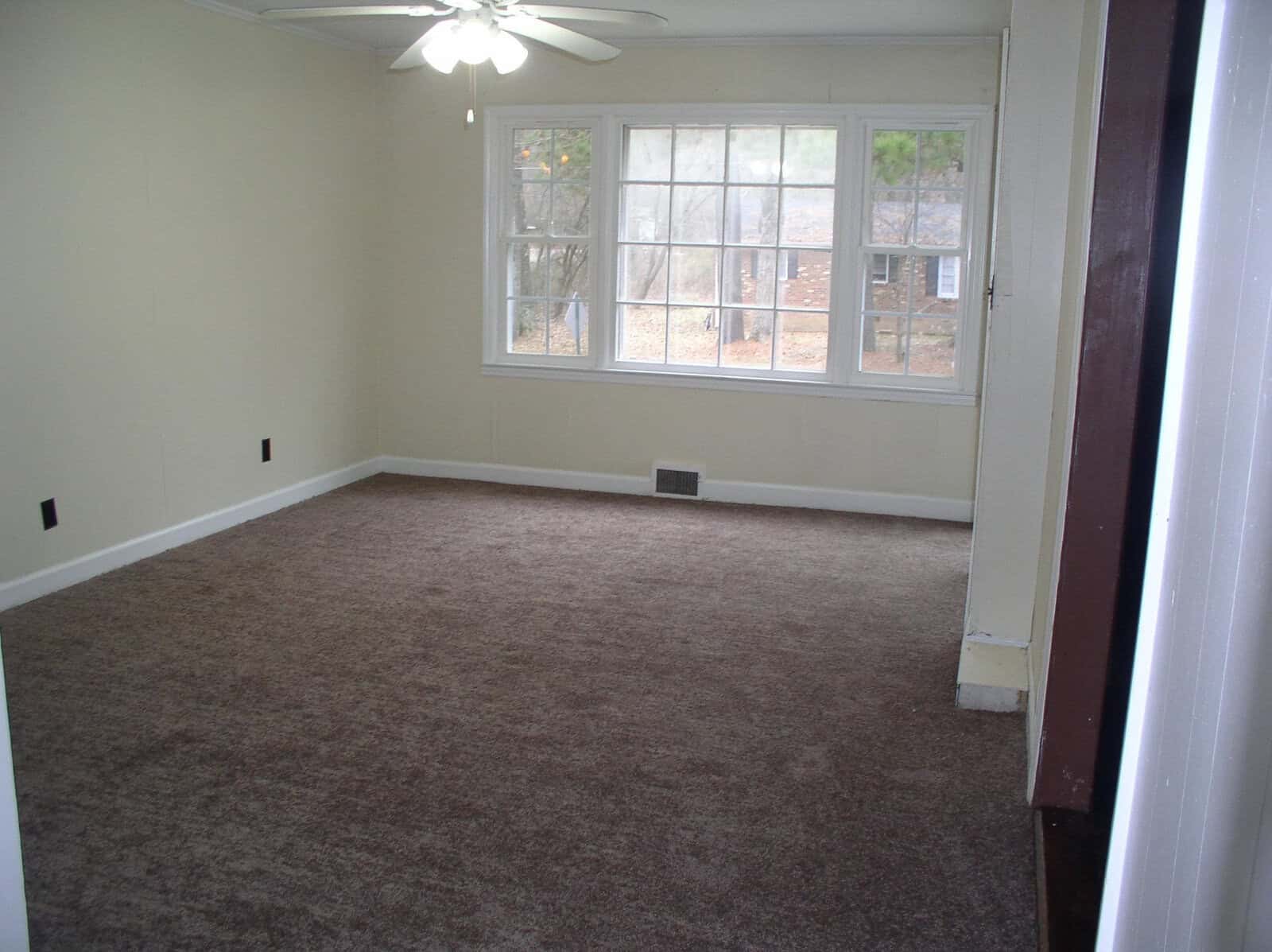 An empty room with beige walls, brown carpet, and a window providing a view of trees outside.