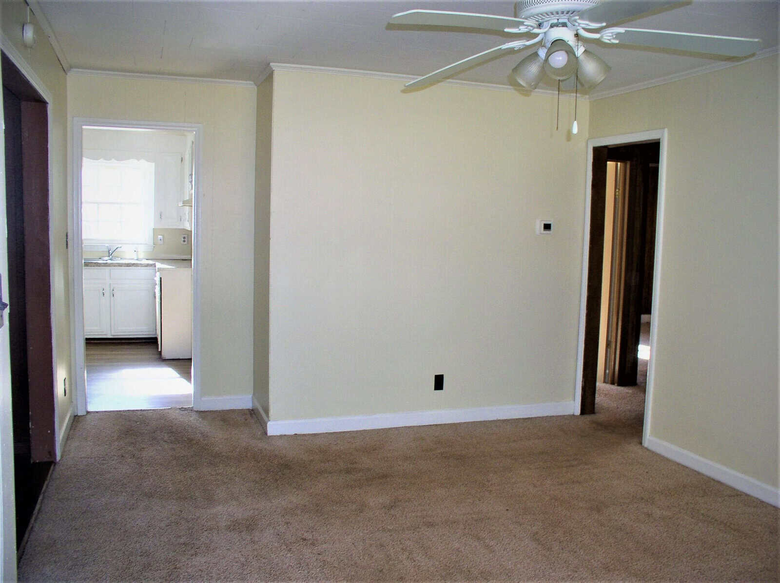 An empty room with beige walls and brown carpet, featuring a ceiling fan, with an open doorway leading to a white kitchen.