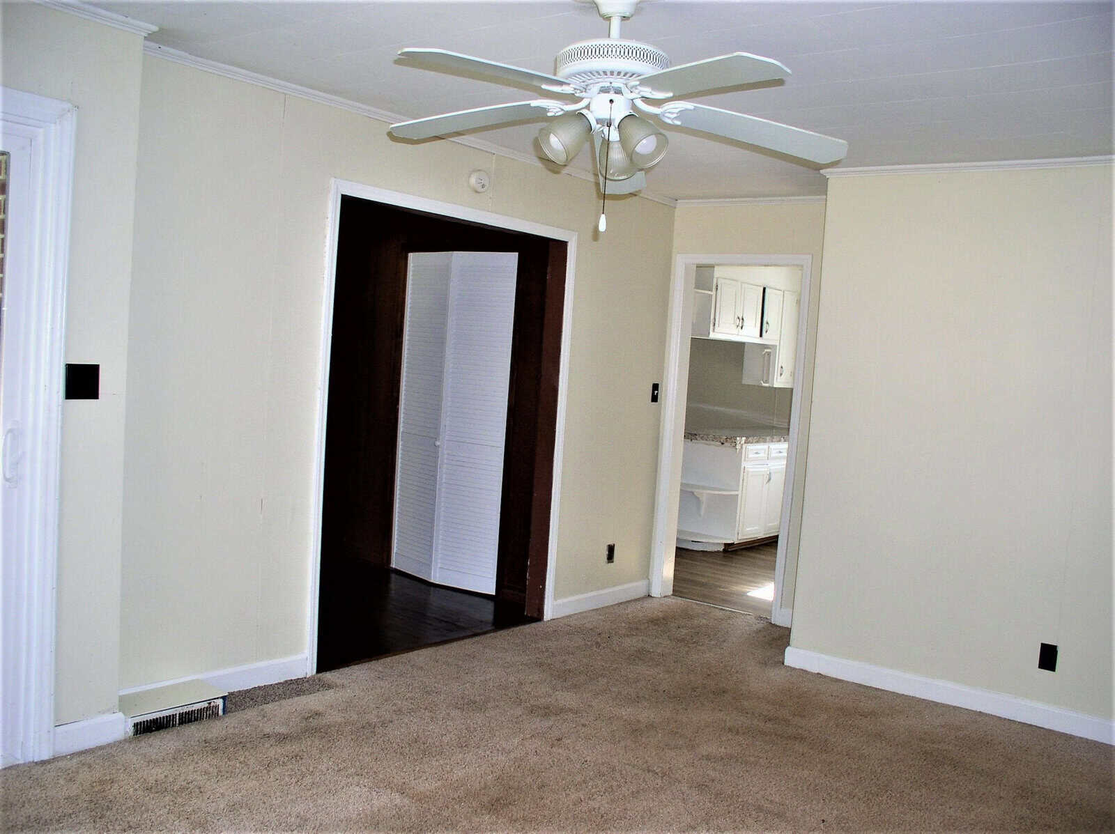 An empty room with beige walls, brown carpet, and a white ceiling fan, featuring an open doorway leading to another room with a partial view of a white kitchen cabinet.