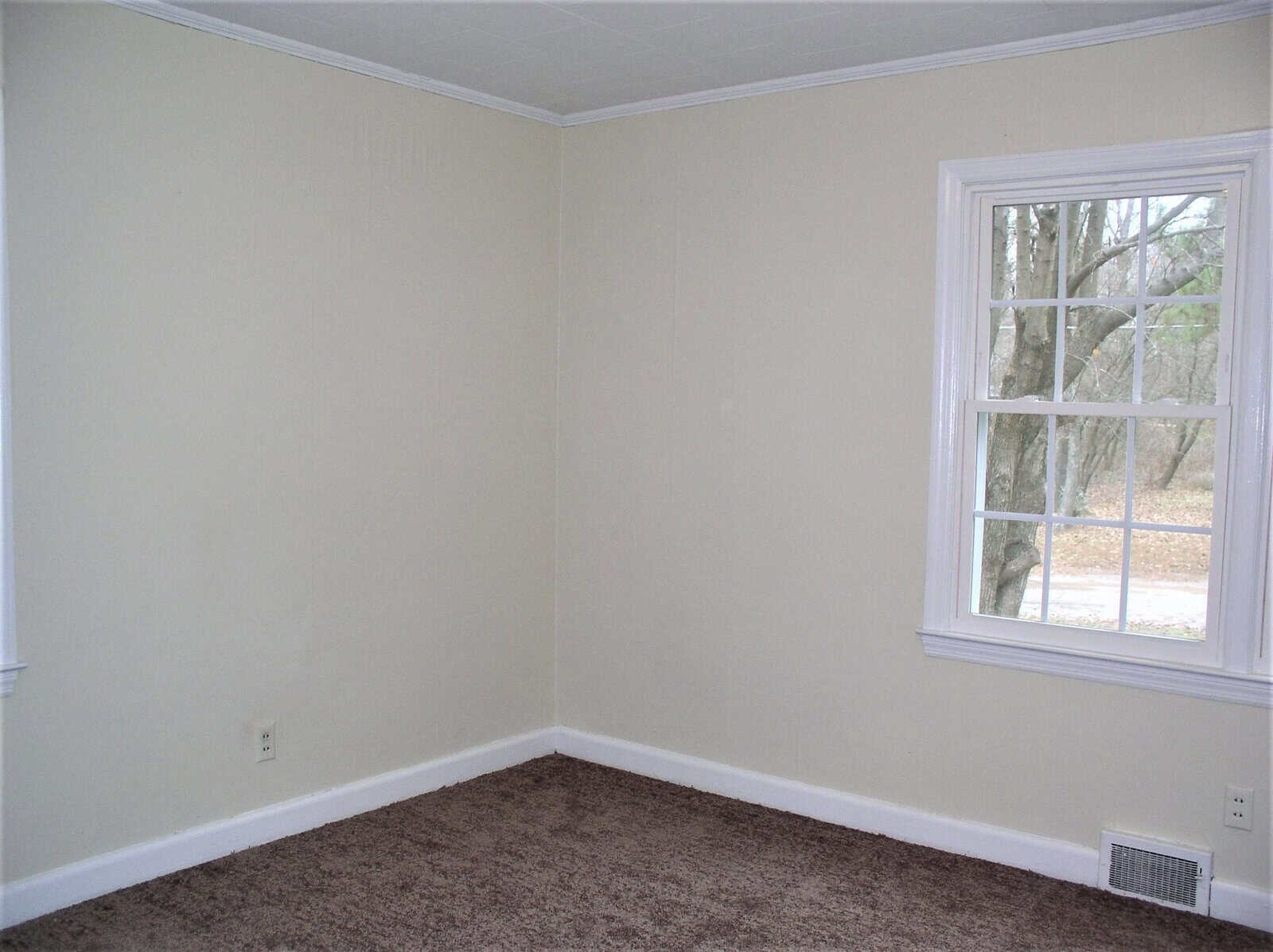 Empty room with beige walls, a white window frame, brown carpet, and a single vent on the floor.