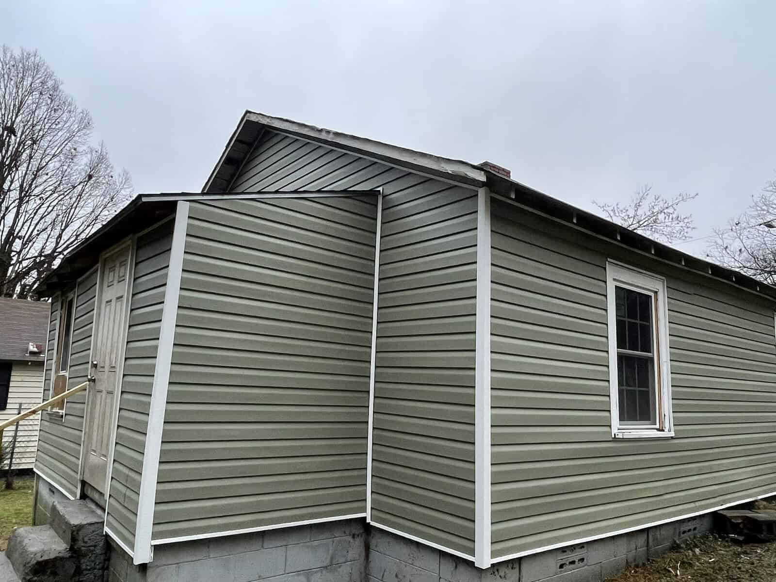 A small, single-story house with olive green siding, a white-trimmed window, and a gabled roof, seen on an overcast day.