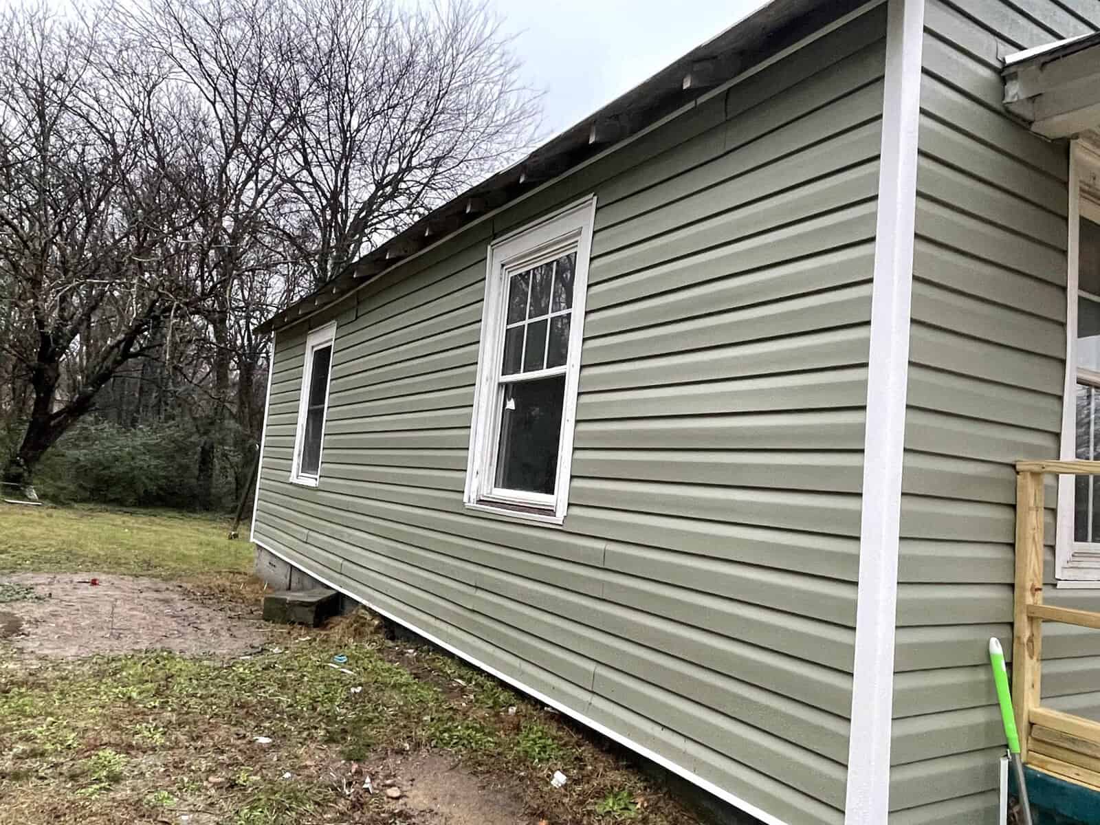 A side view of a green single-story house with white trim, featuring two windows and a partial view of a porch with a railing. the ground appears wet, possibly from recent rain, and there are leafless trees in the background, suggesting it might be late fall or winter.