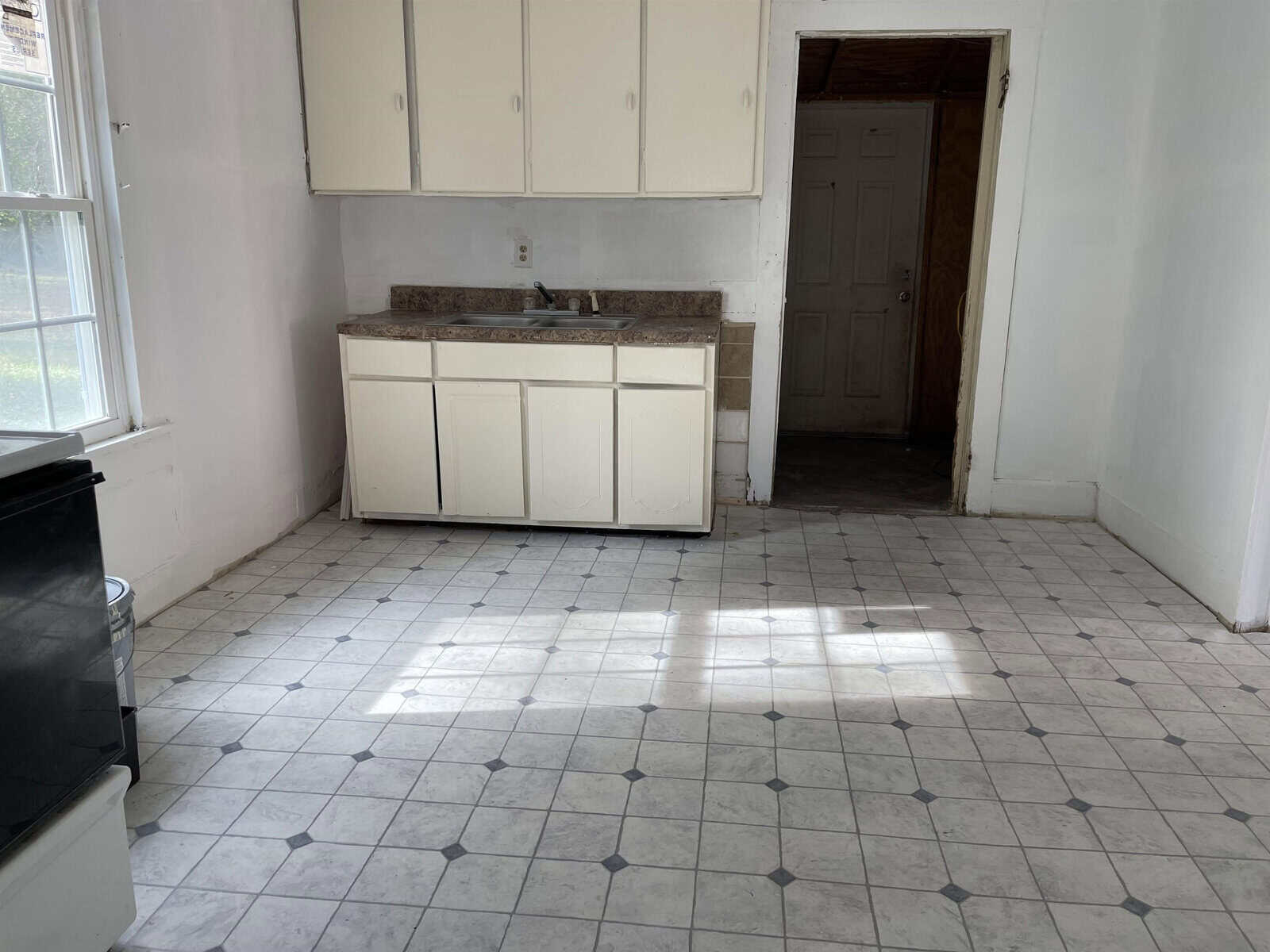 A sparsely furnished kitchen with white cabinets, a countertop with a sink, a stove on the left, and tiled flooring, with natural light coming through the window.
